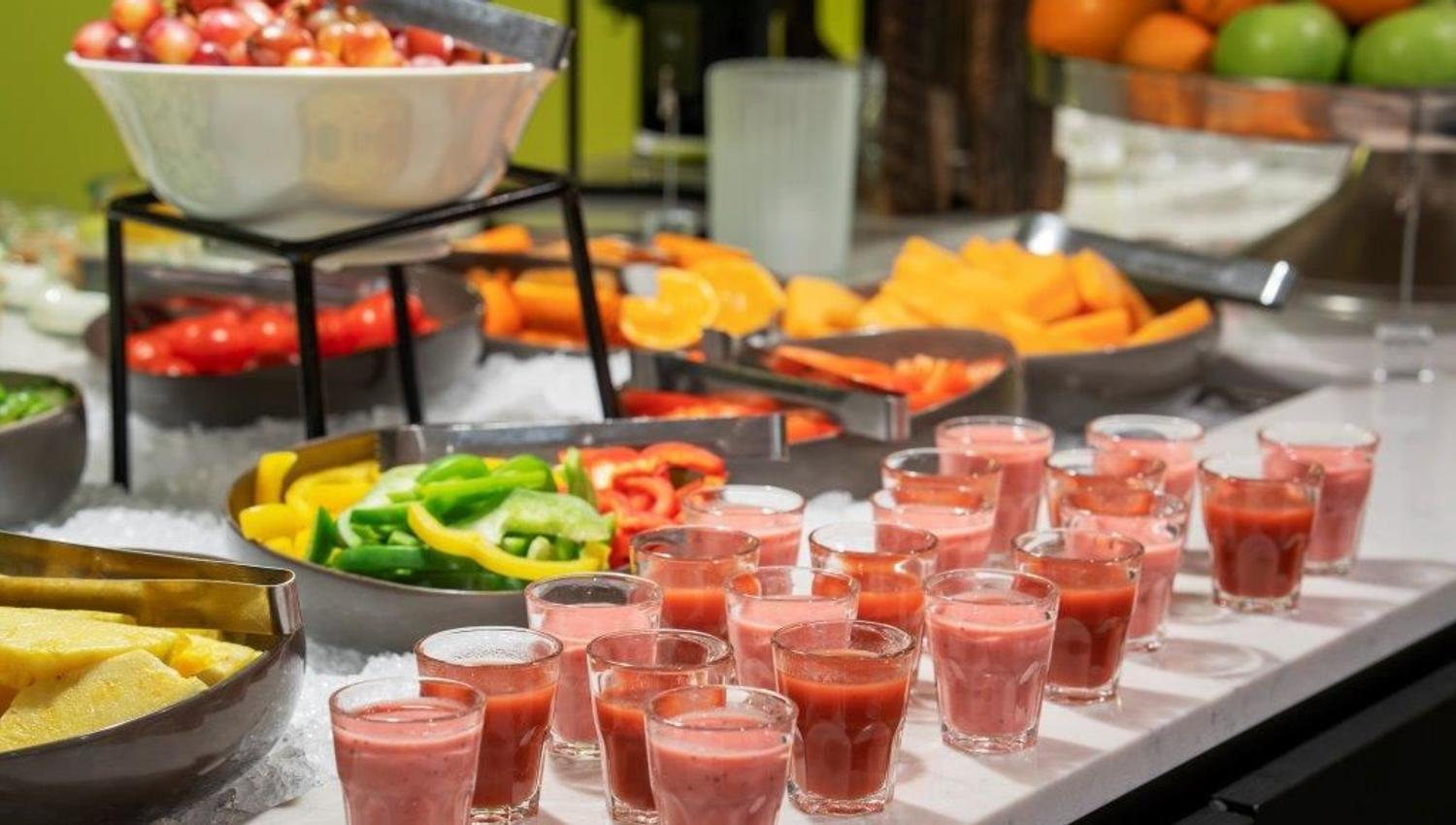 Buffet table with fruit, vegetables, and small glasses of pink smoothie.