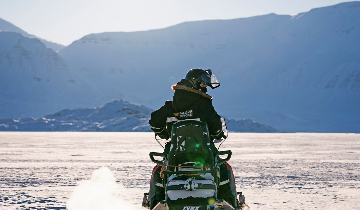 A guest wearing snowmobile equipment sitting on a snowmobile in a snow-covered landscape while looking behind themselves 
