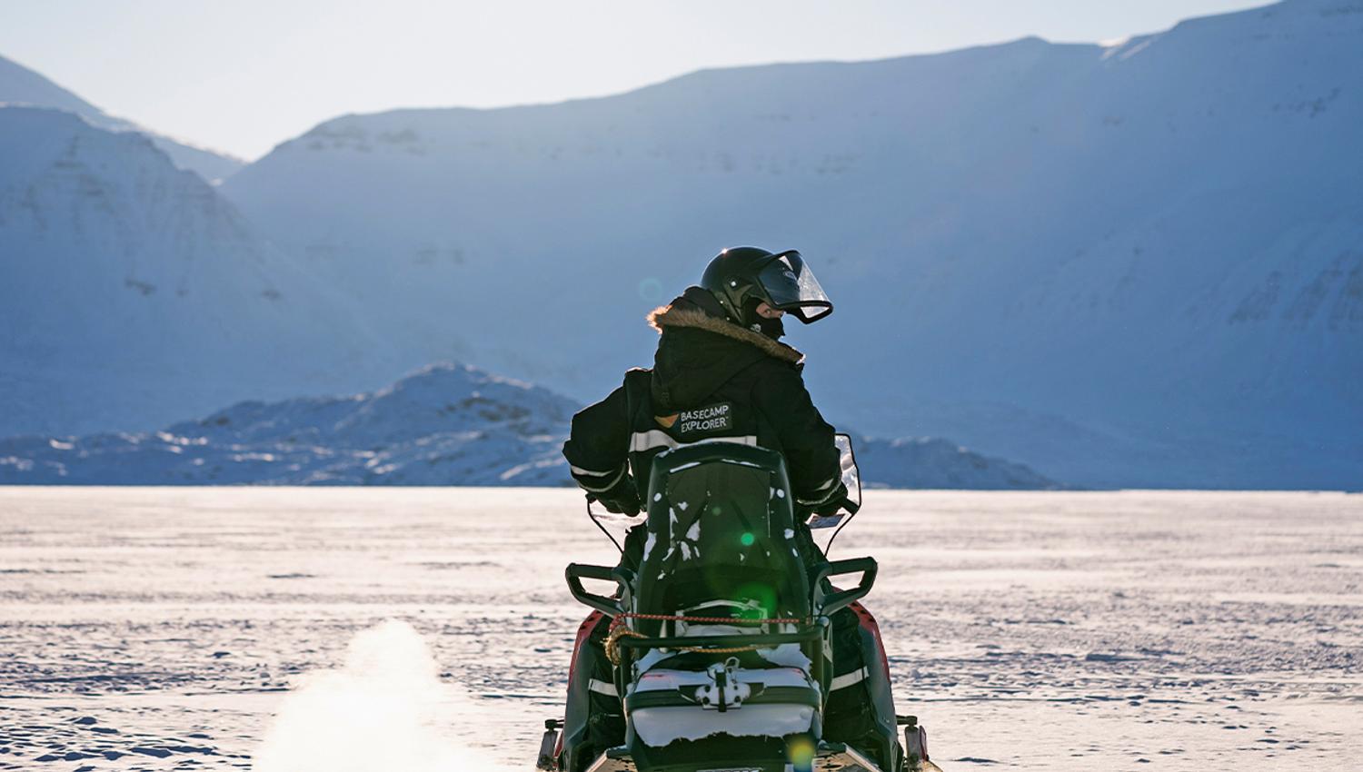 A guest wearing snowmobile equipment sitting on a snowmobile in a snow-covered landscape while looking behind themselves
