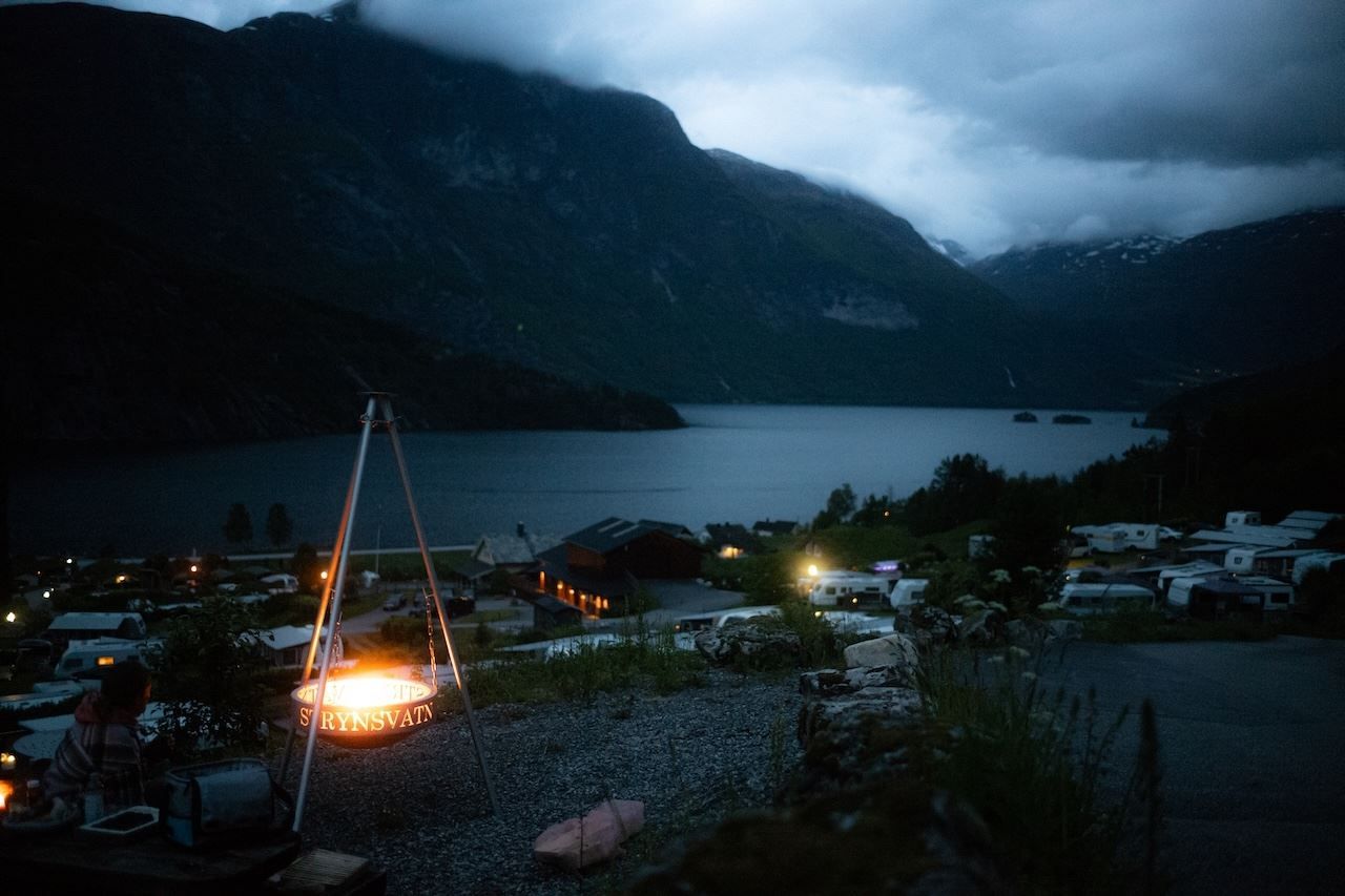 A glowing campfire in a tripod brazier overlooks a lakeside village and dark mountains beneath cloudy dusk skies..
