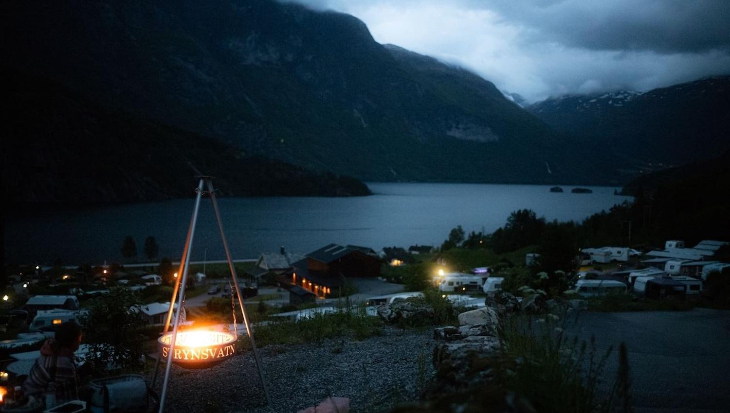 A glowing campfire in a tripod brazier overlooks a lakeside village and dark mountains beneath cloudy dusk skies..