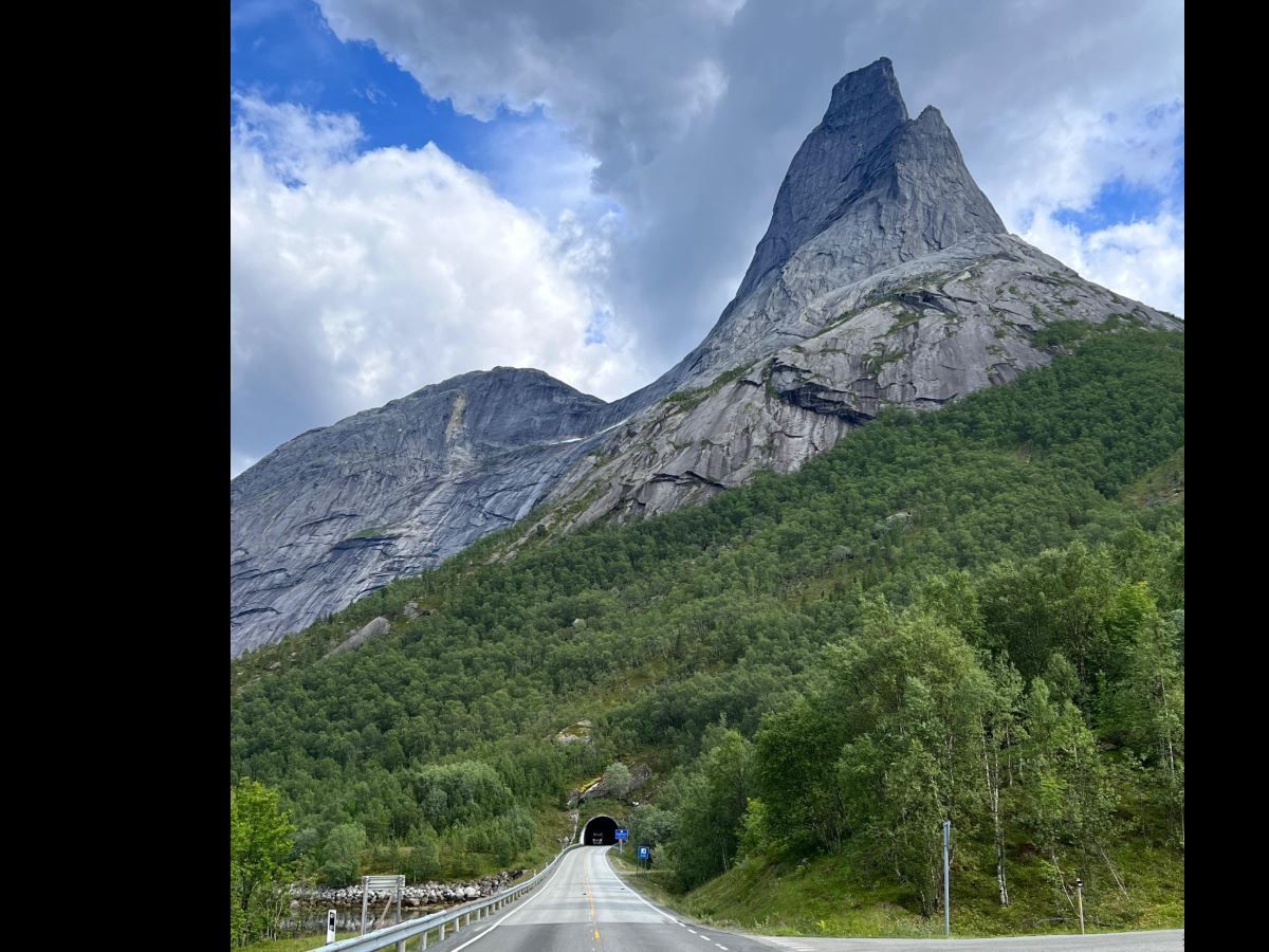 En asfaltert vei leder inn i en tunnel ved foten av et bratt, grått fjell med grønn skog og skyet himmel..