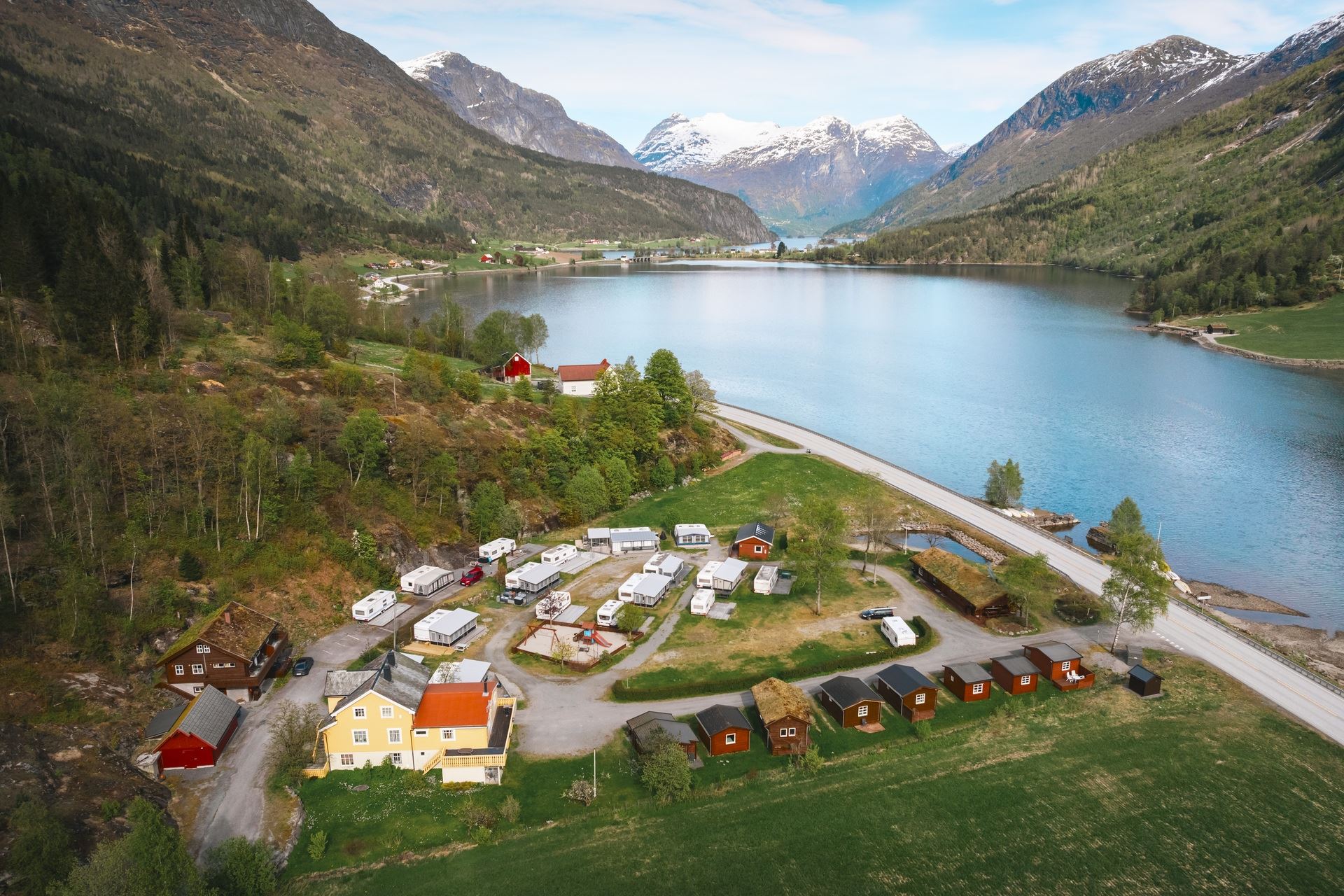 A small lakeside village with cabins and caravans sits beside a blue lake, surrounded by green mountains and snowy peaks..