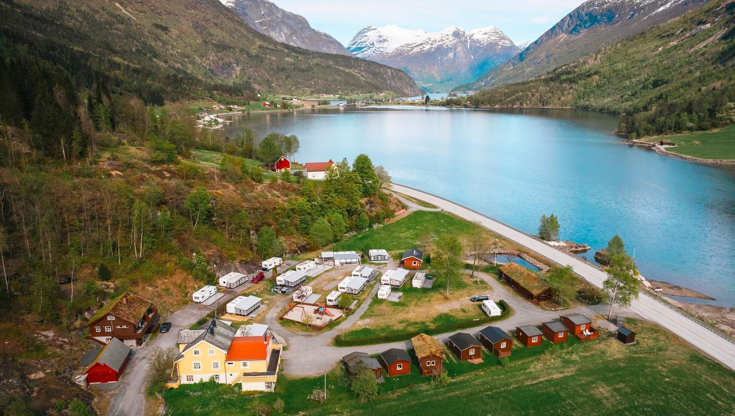 A small lakeside village with cabins and caravans sits beside a blue lake, surrounded by green mountains and snowy peaks..