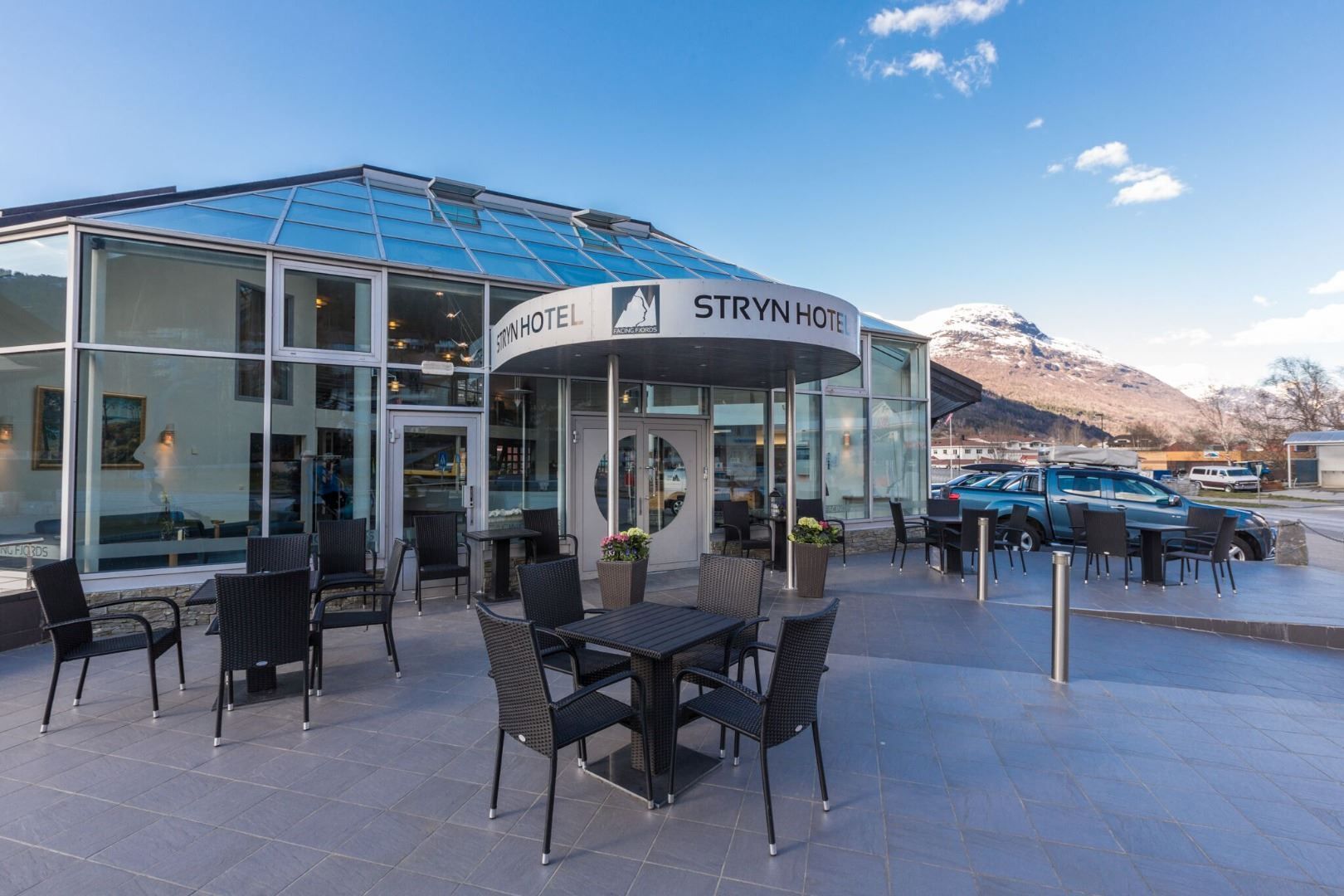 Glasfasade på hotell med utebord og stolar på terrasse, parkerte bilar og snødekt fjell under blå himmel..