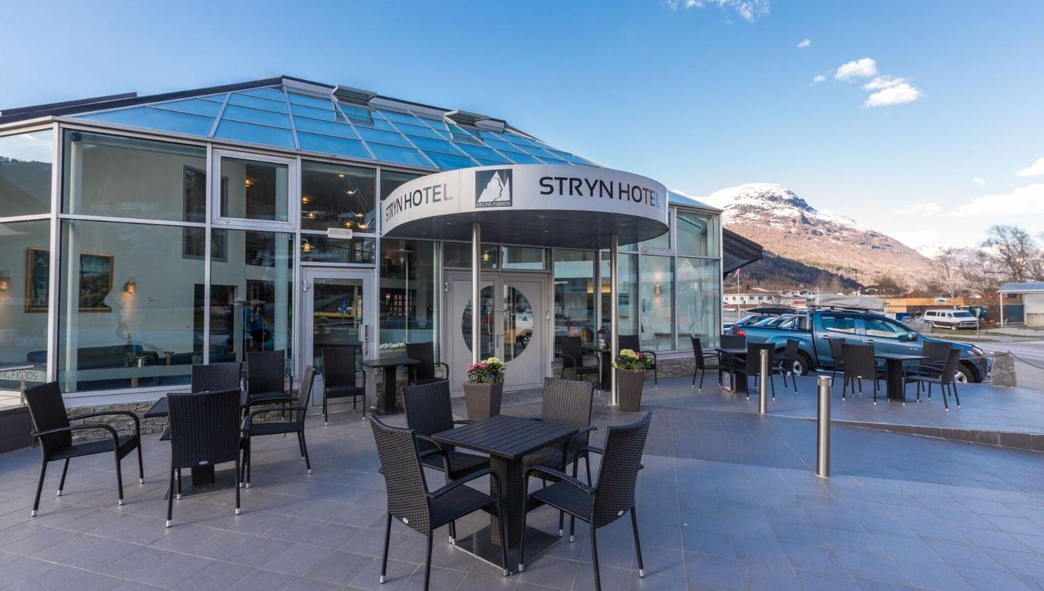 Glasfasade på hotell med utebord og stolar på terrasse, parkerte bilar og snødekt fjell under blå himmel..