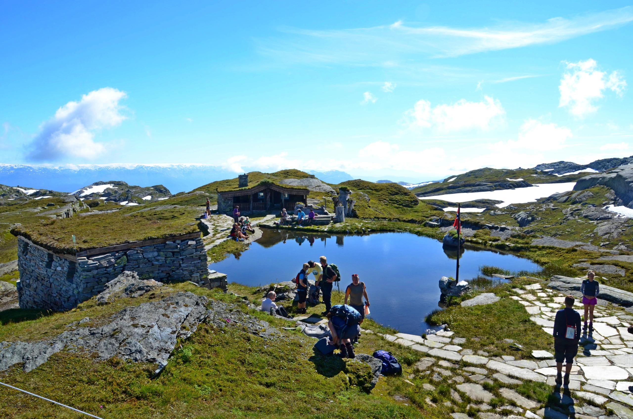 Hikers relaxing at Breidablik mountain lodge beside a small alpine lake in Fitjadalen, surrounded by mountains and patches of snow.
