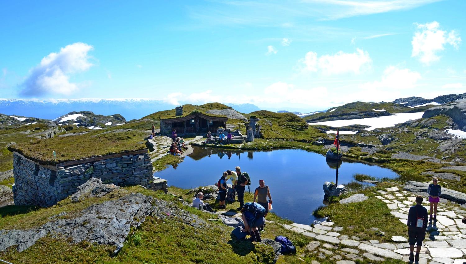 Hikers relaxing at Breidablik mountain lodge beside a small alpine lake in Fitjadalen, surrounded by mountains and patches of snow.