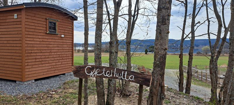 Wooden sign reading Gjeterhytta next to a newly built mini cabin surrounded by trees and open landscape