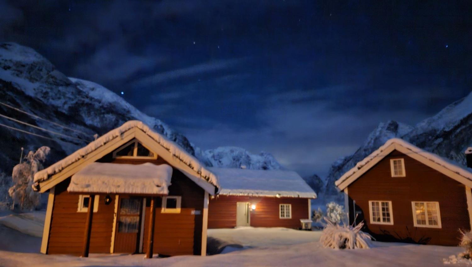 Three snow-covered wooden cabins lit by warm lights sit in a winter landscape beneath a dark, starry sky, at Aabrekk Gard Trollbu in Oldedalen valley.