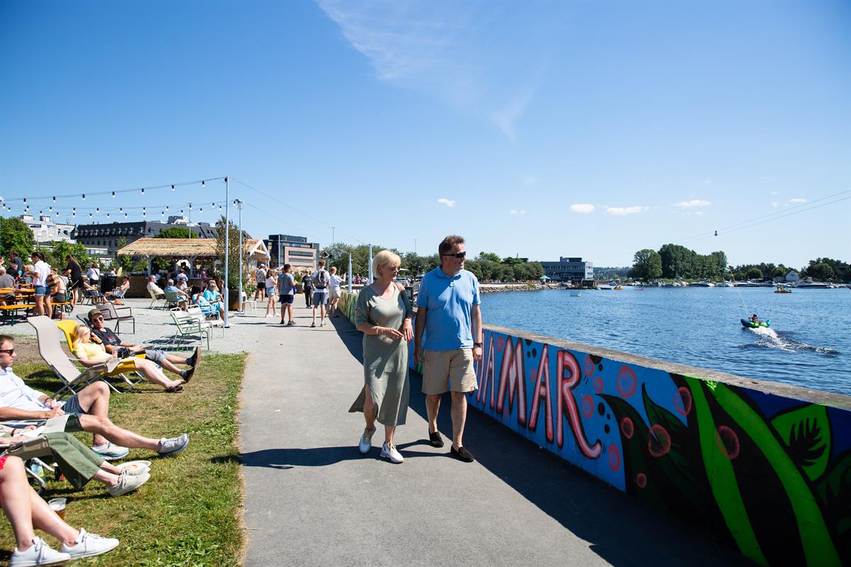 Folk som sitter og går langs vannkanten ved en fargerik veggmaleri-installasjon.