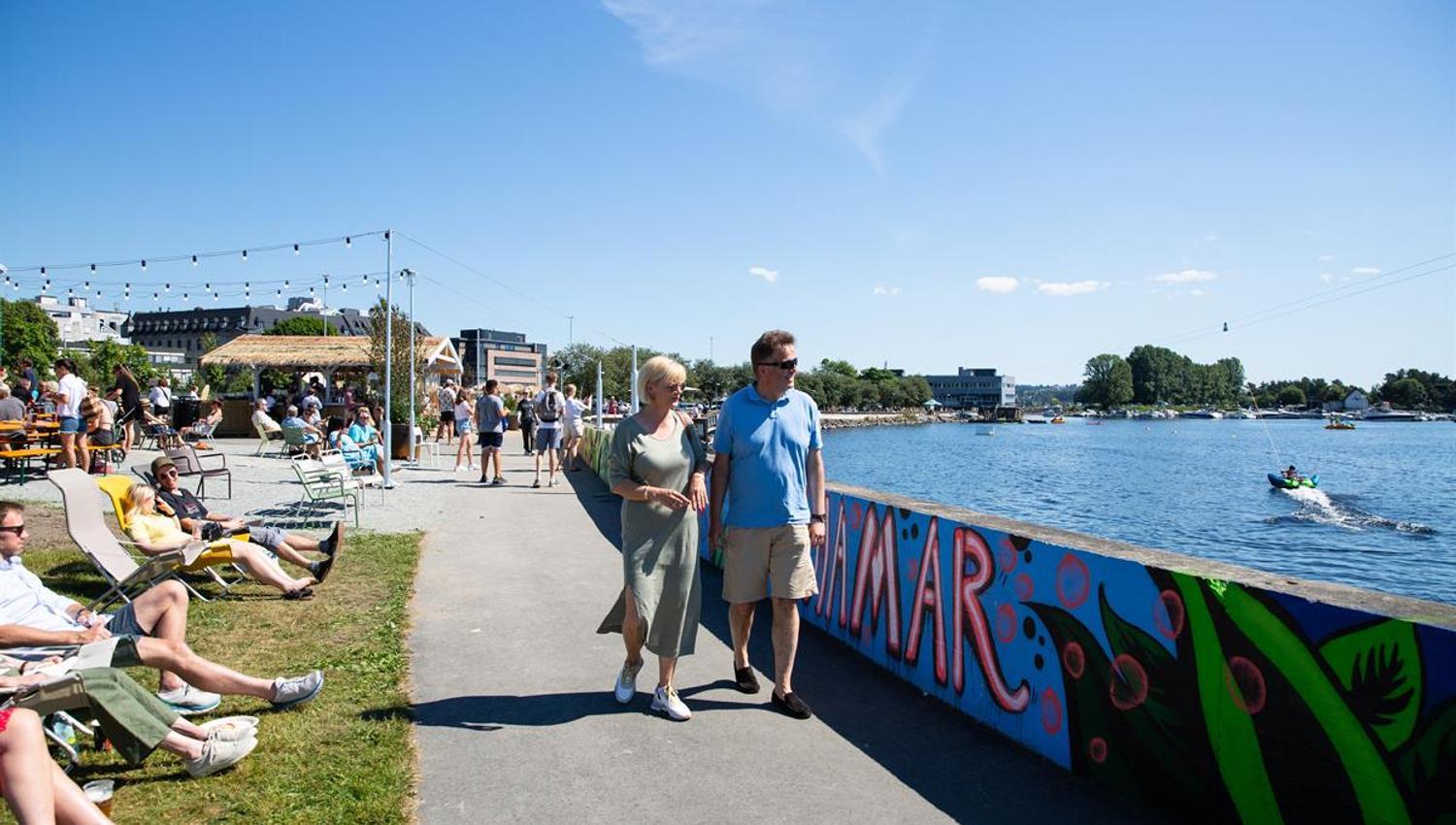 Folk som sitter og går langs vannkanten ved en fargerik veggmaleri-installasjon.