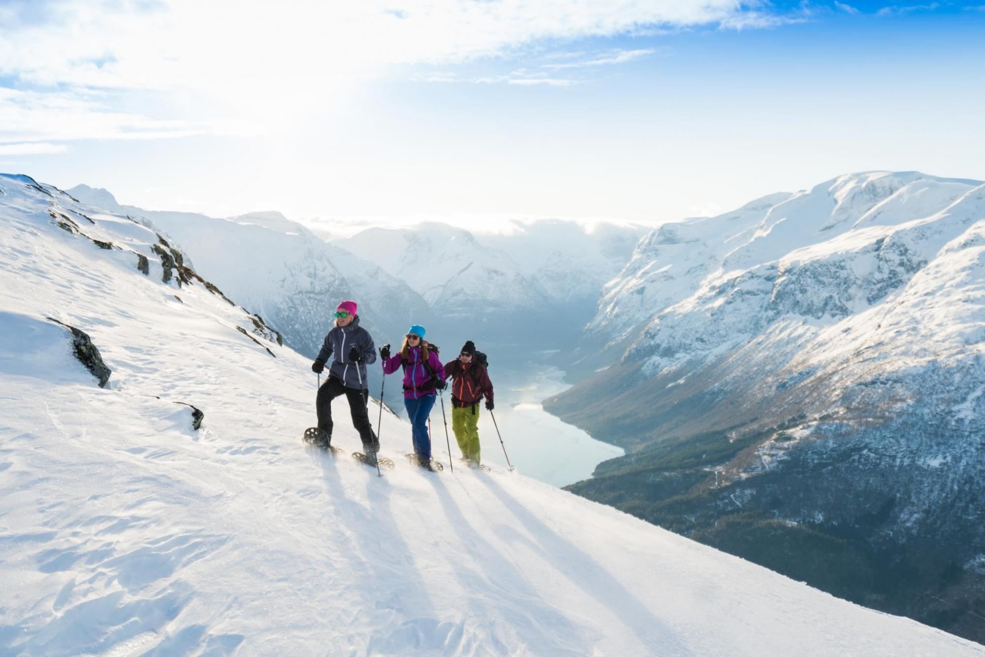 Three hikers in Loen with backpacks and trekking poles snowshoe across a sunlit slope above a fjord and snowy peaks..
