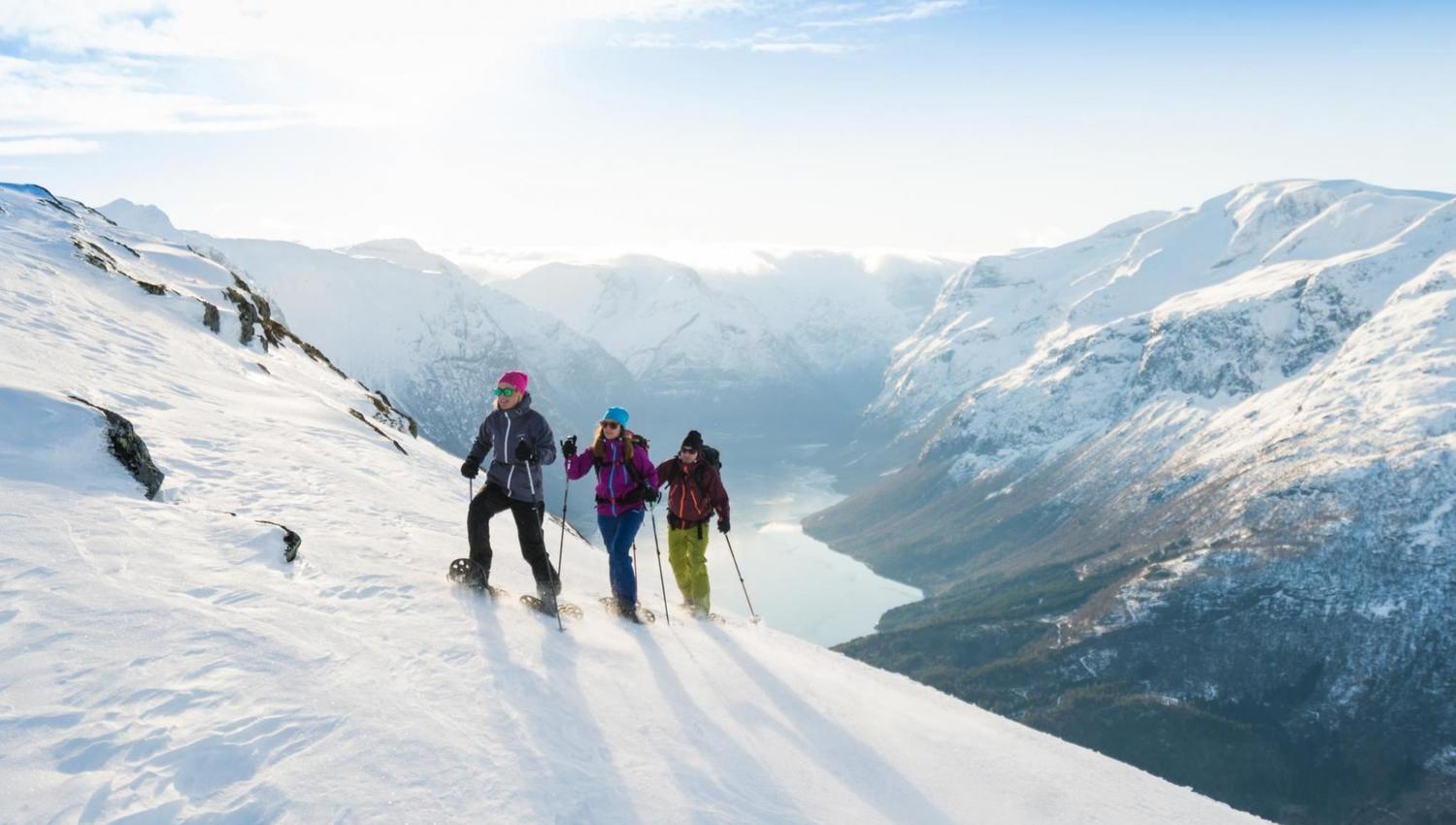Three hikers in Loen with backpacks and trekking poles snowshoe across a sunlit slope above a fjord and snowy peaks..