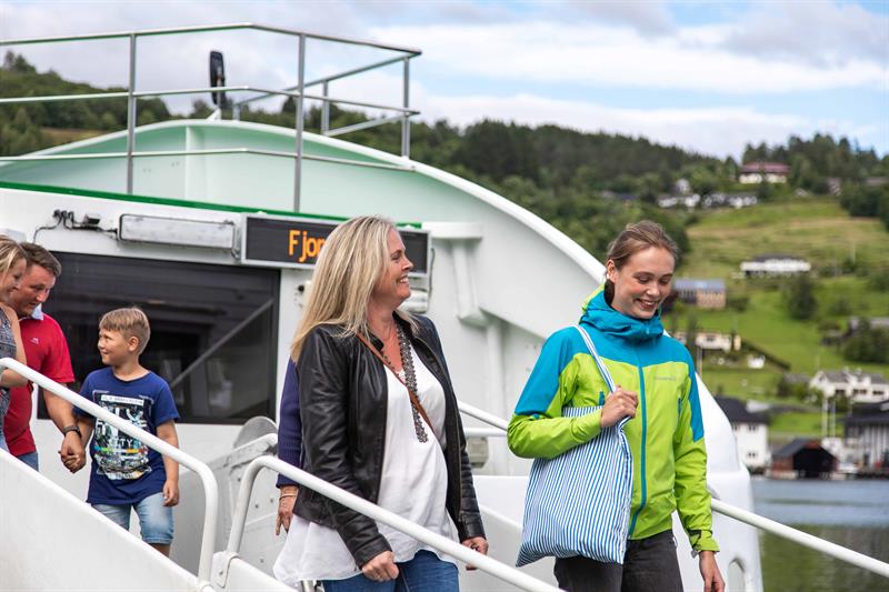 Passasjerar går i land frå Norled-cruisebåten ved ein fjordkai i Hardanger, grøne lier i bakgrunnen.