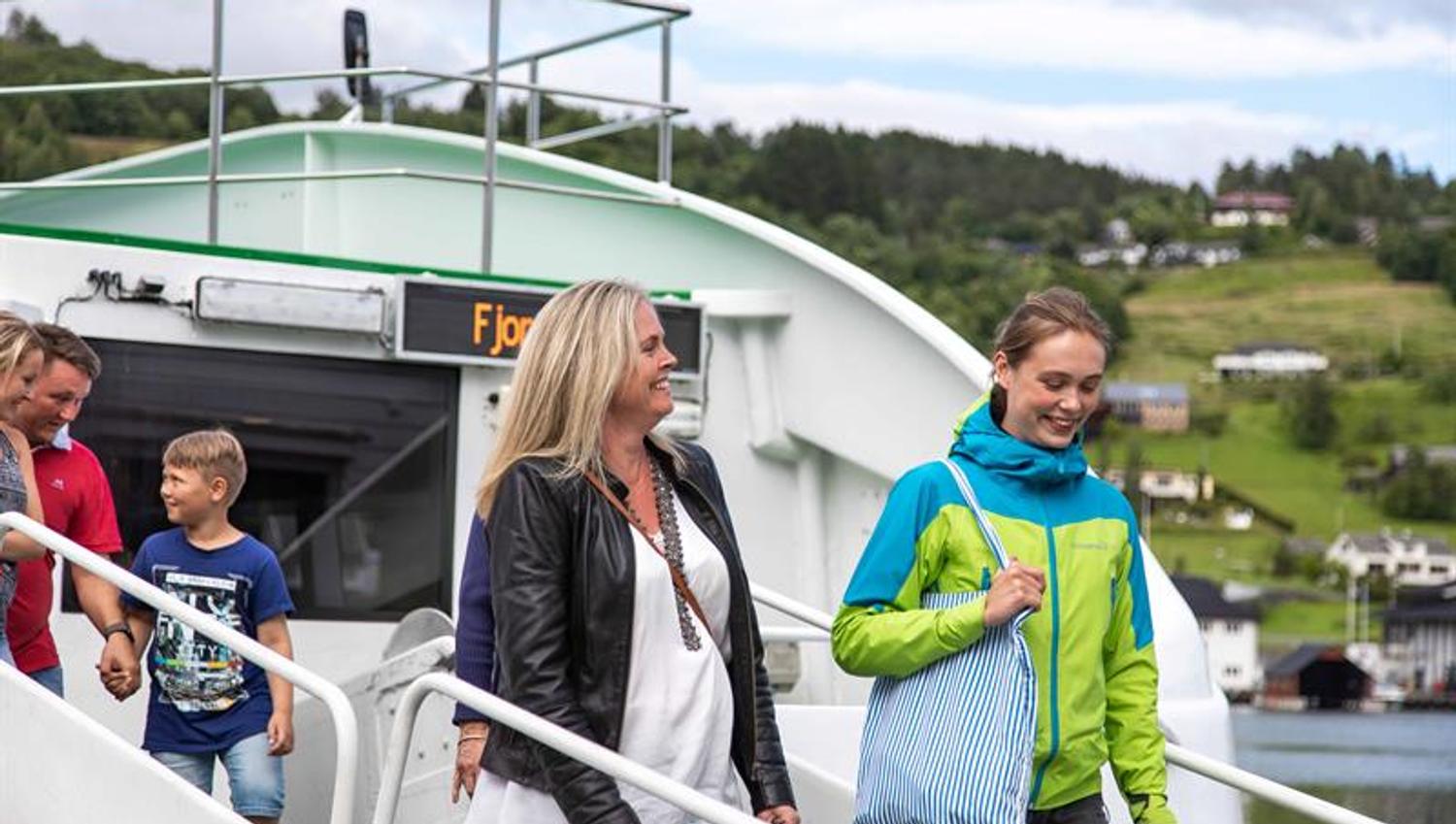 Passasjerar går i land frå Norled-cruisebåten ved ein fjordkai i Hardanger, grøne lier i bakgrunnen.