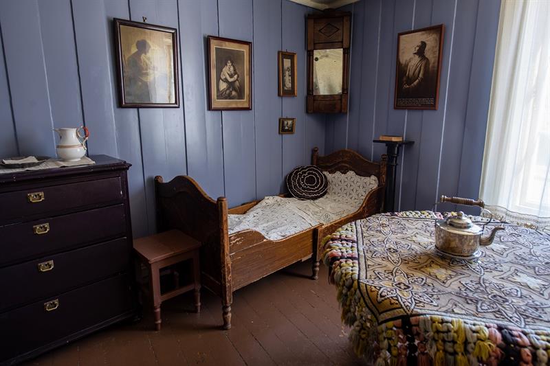 Historic interior featuring a single bed, chest of drawers, table and framed pictures on blue-painted walls.