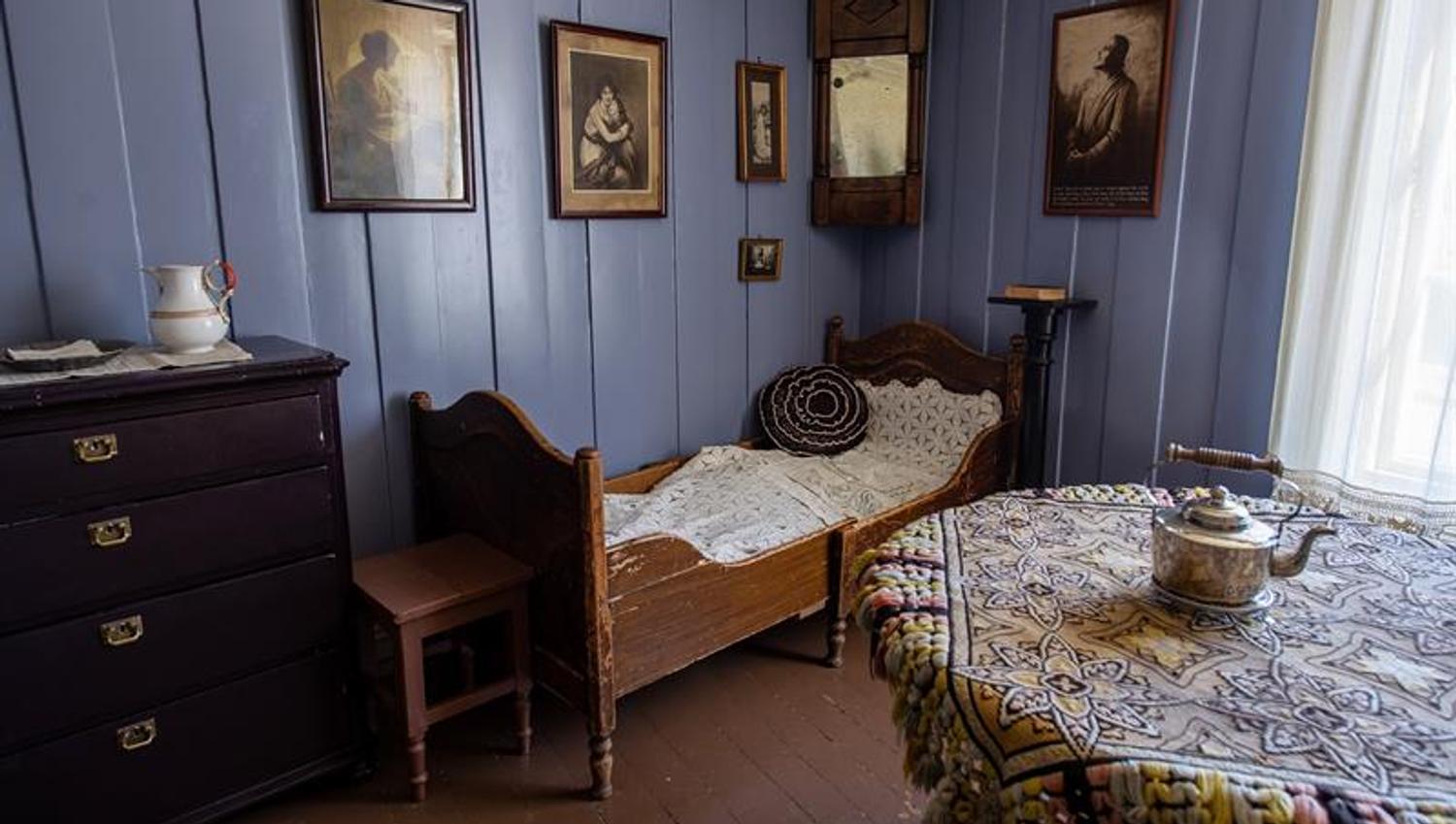 Historic interior featuring a single bed, chest of drawers, table and framed pictures on blue-painted walls.