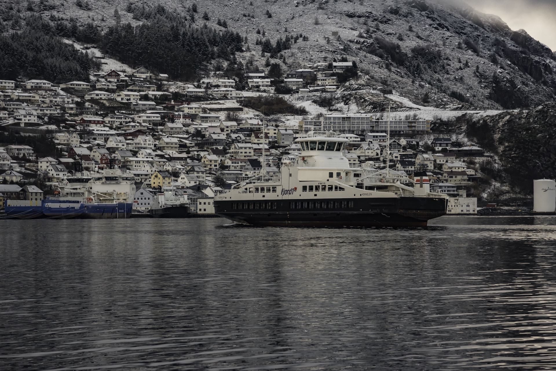 A largeFjord1  ferry crosses calm water in front of a hillside town with snow-dusted roofs and mountains..