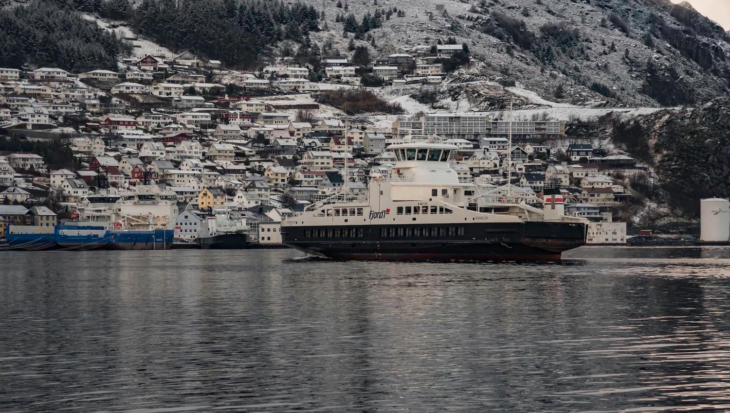 A largeFjord1 ferry crosses calm water in front of a hillside town with snow-dusted roofs and mountains..