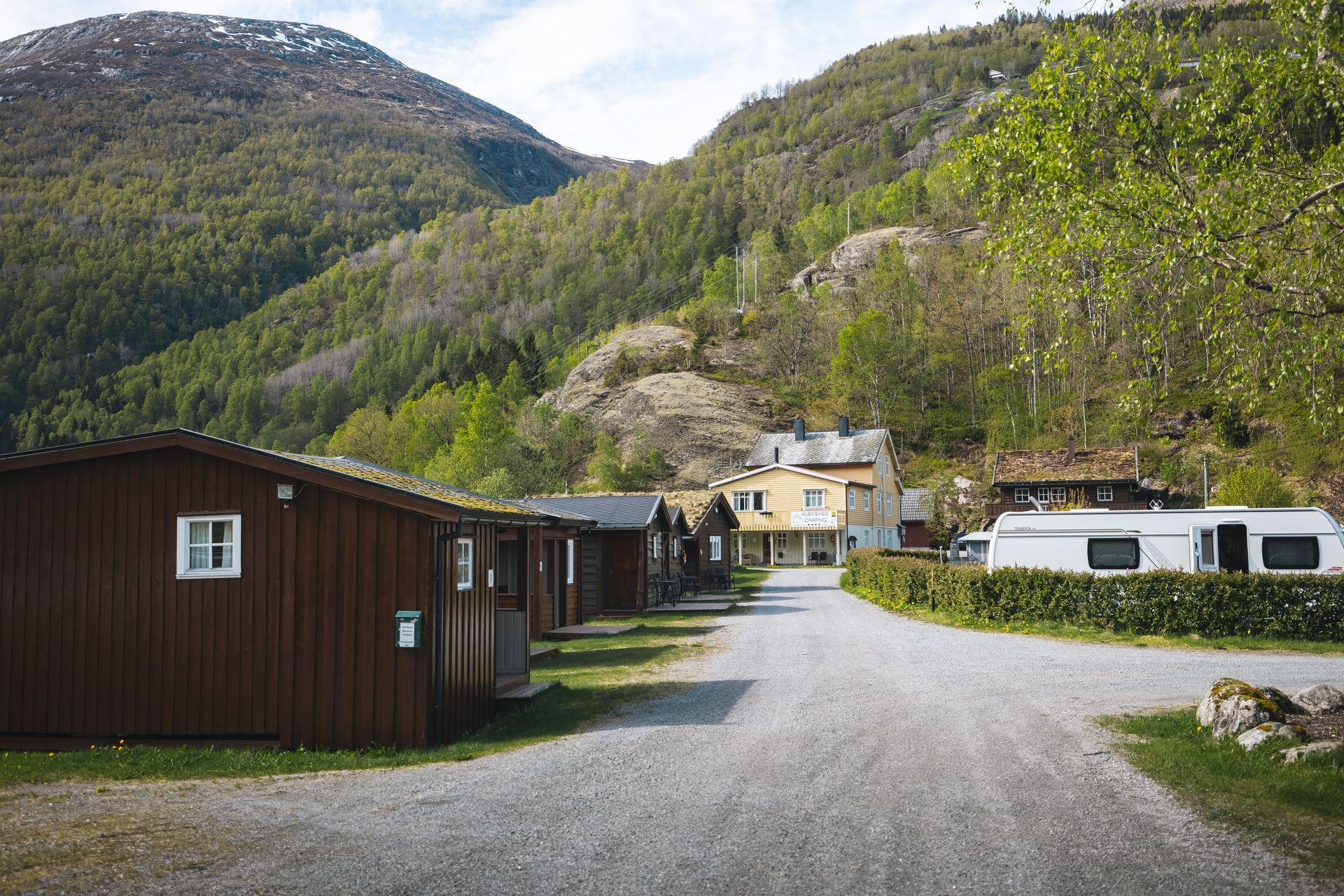 Gravel road past wooden cabins and a camper, leading to a small building beneath green forested mountains..