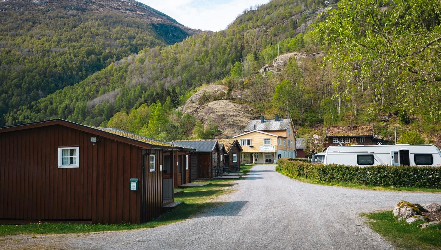 Gravel road past wooden cabins and a camper, leading to a small building beneath green forested mountains..