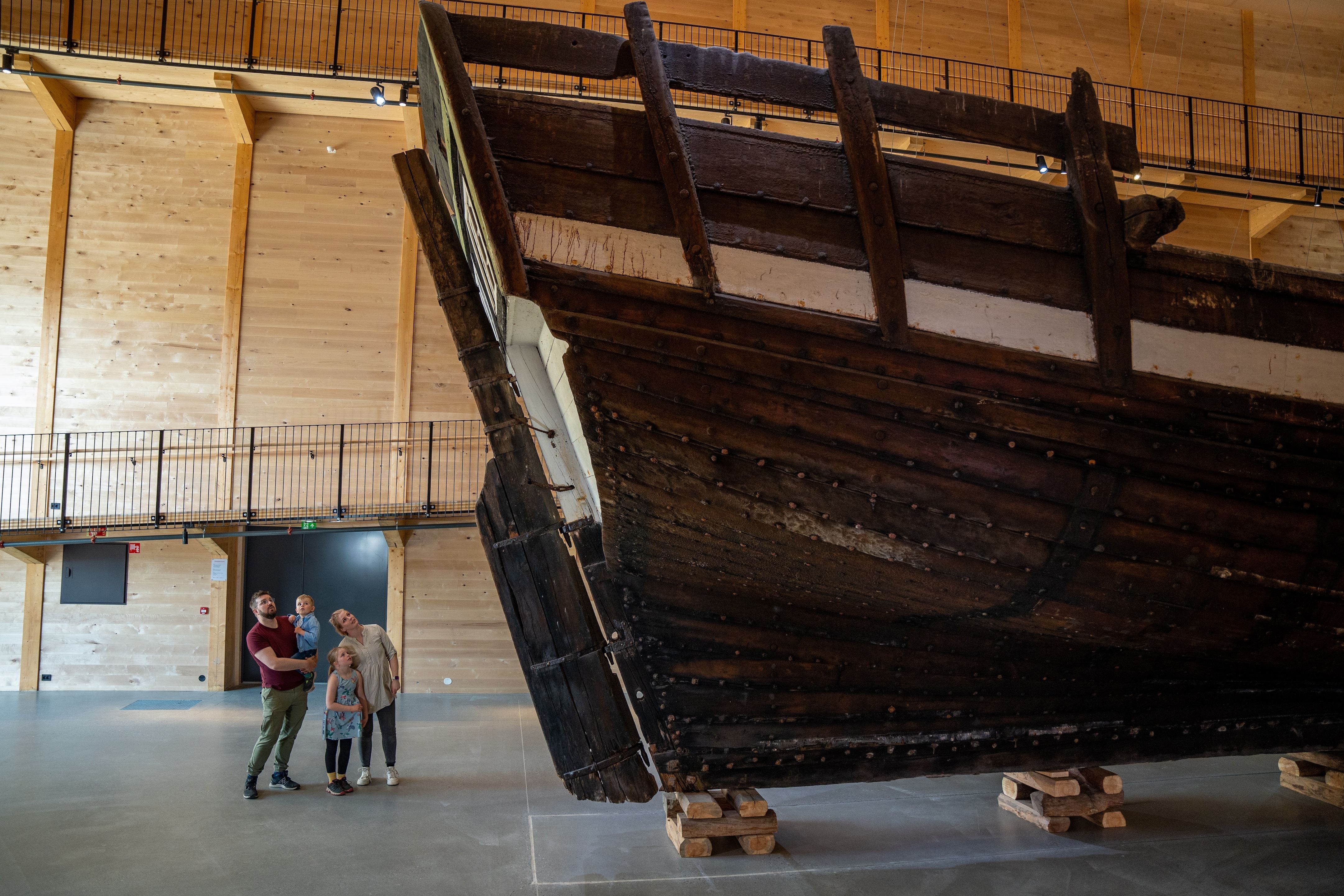 Large dark wooden ship hull displayed indoors on timber supports, with visitors nearby in a spacious hall..