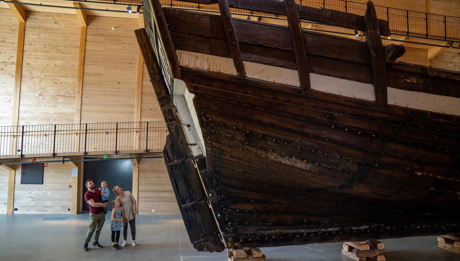 Large dark wooden ship hull displayed indoors on timber supports, with visitors nearby in a spacious hall..