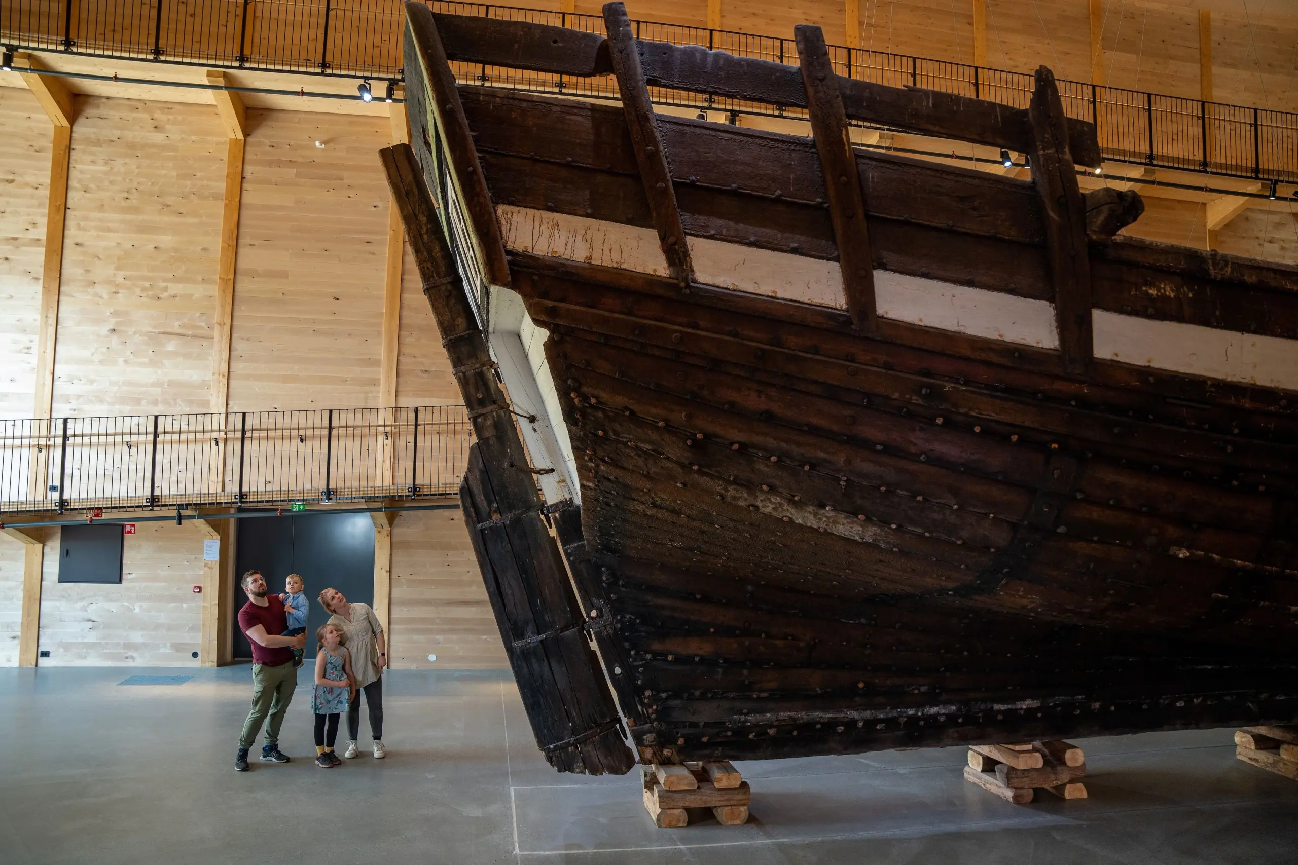 Large dark wooden ship hull displayed indoors on timber supports, with visitors nearby in a spacious hall..
