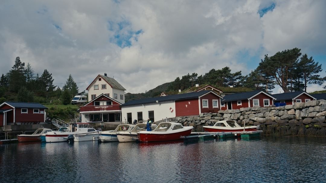 Small boats moored at a quiet marina beside red wooden buildings and a rocky seawall under a cloudy sky..