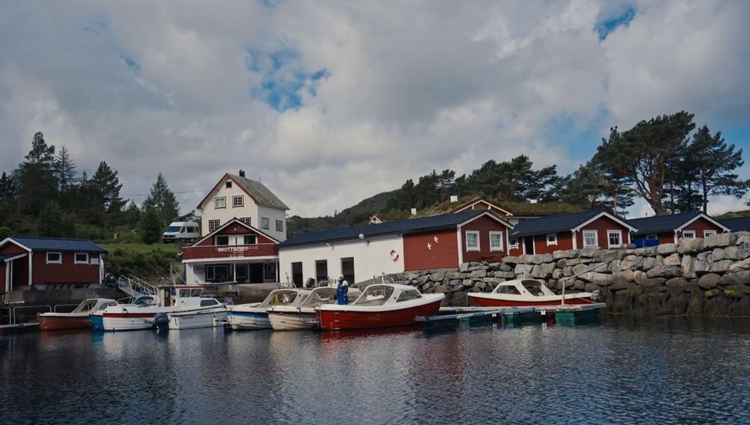 Small boats moored at a quiet marina beside red wooden buildings and a rocky seawall under a cloudy sky..