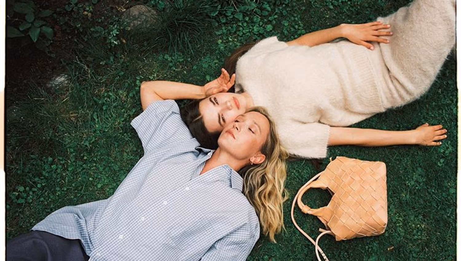 Two girls laying on a lawn, next to a beige handbag.