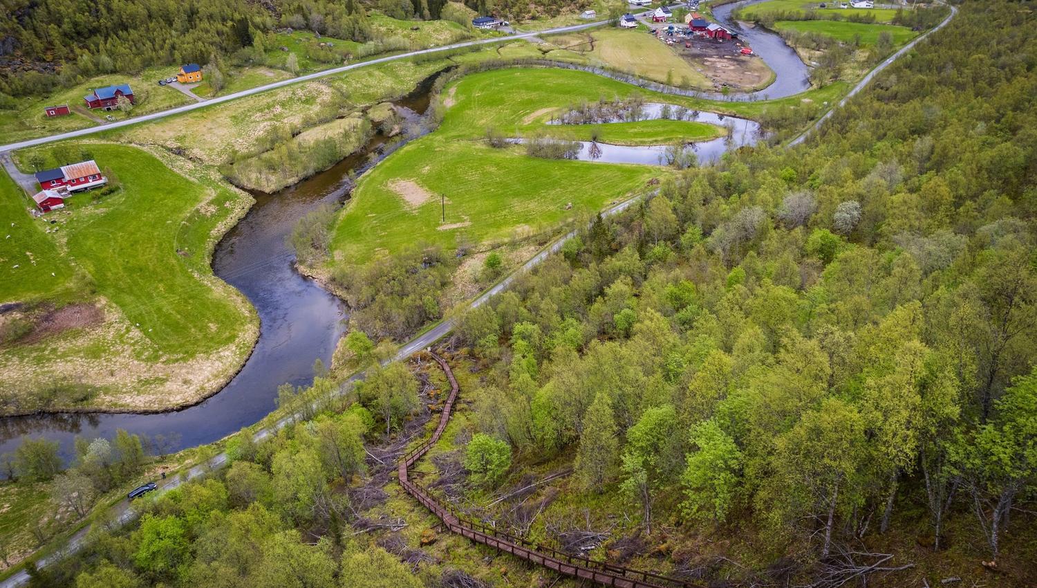 Aerial view of a winding river through green fields and forest, with scattered farmhouses and a road nearby.