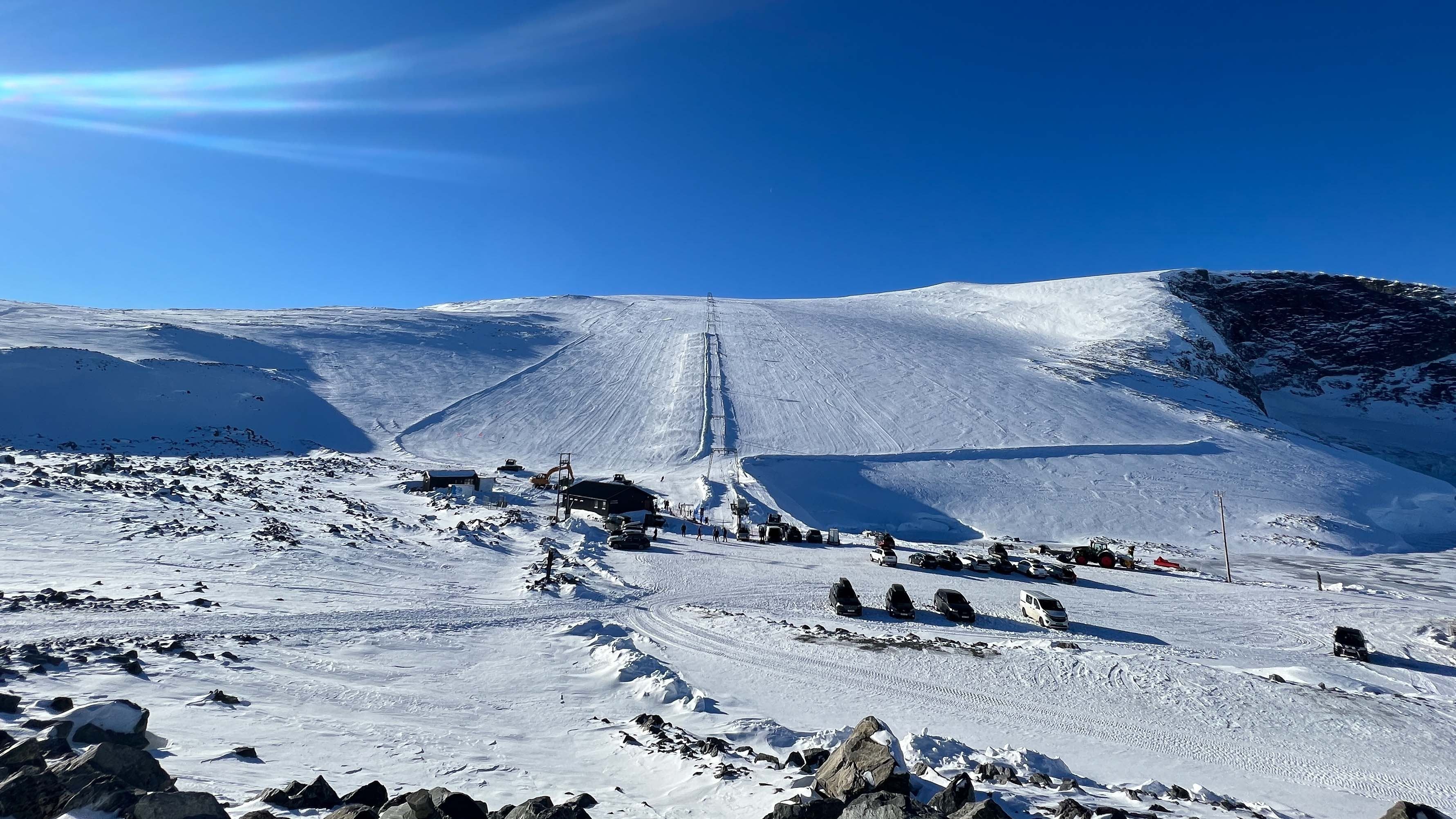 Wide view of a snow-covered mountain slope with a ski lift line, small buildings, and parked vehicles under blue sky..