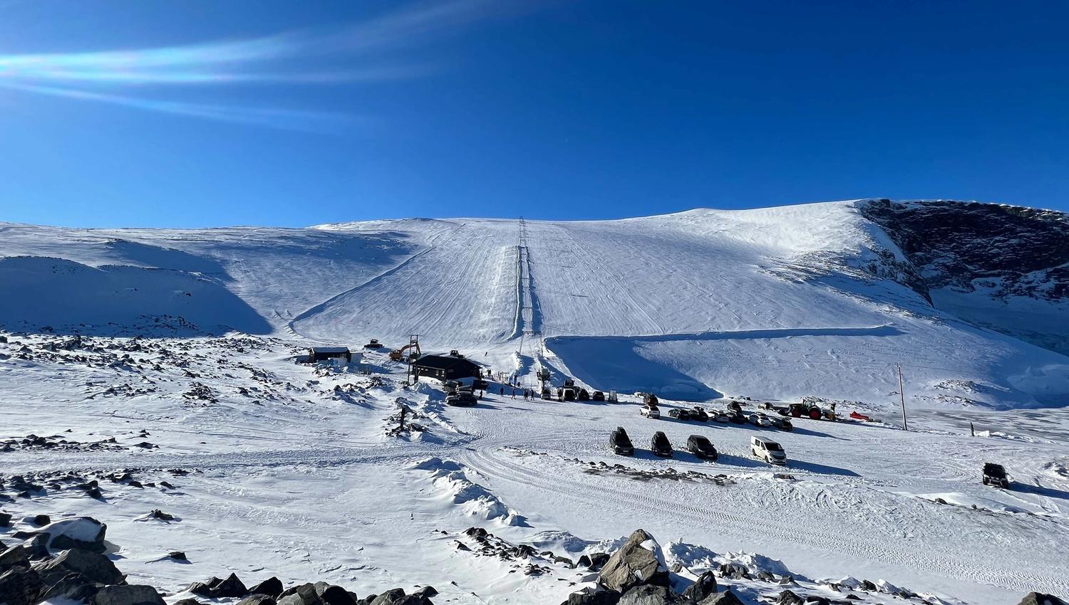 Wide view of a snow-covered mountain slope with a ski lift line, small buildings, and parked vehicles under blue sky..