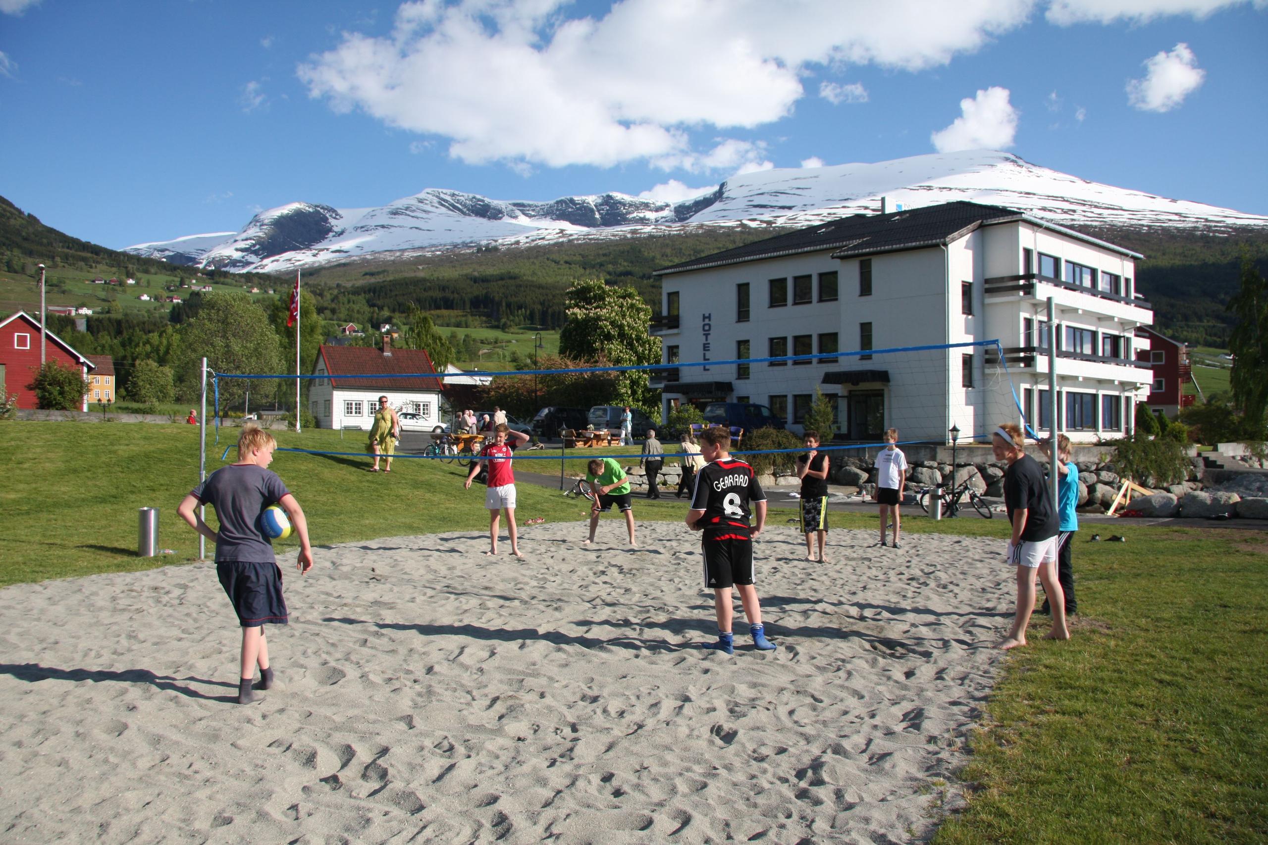 Ei gruppe ungdommar speler sandvolleyball på ei bane ved eit hotell, med snødekte fjell i bakgrunnen..