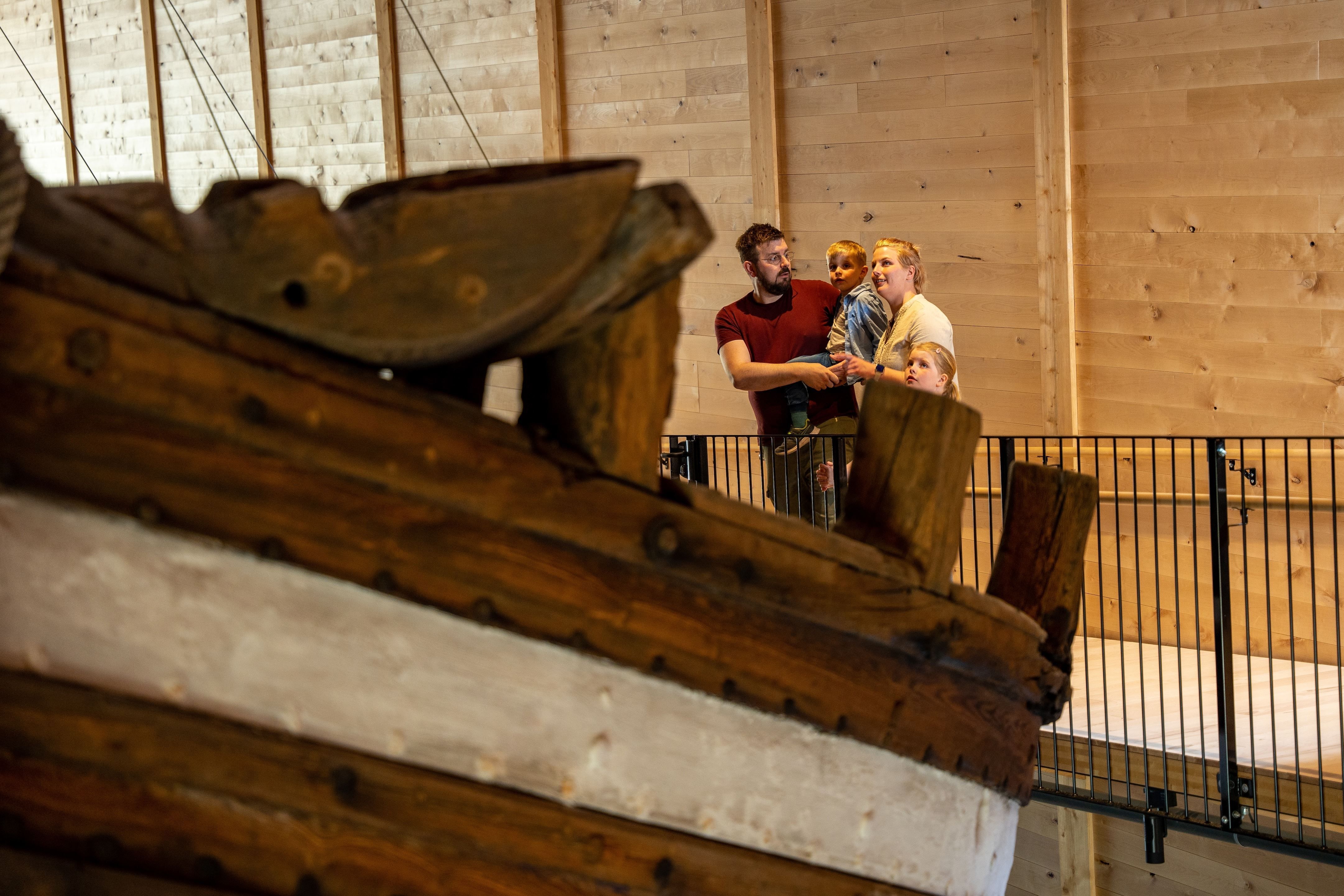 Large wooden ship structure in the foreground with three people standing by a railing inside a timber-walled hall..
