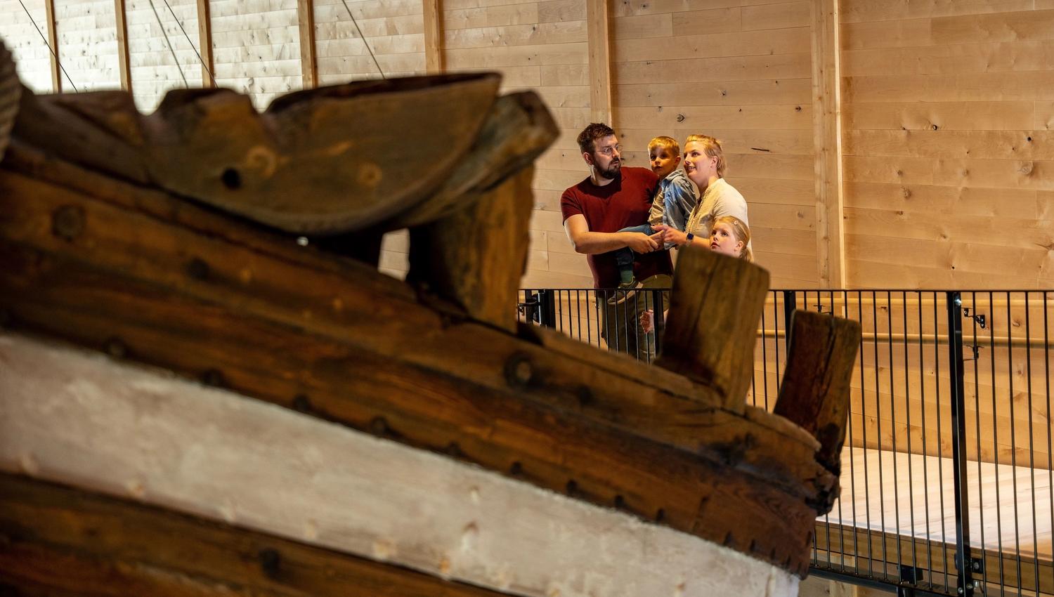 Large wooden ship structure in the foreground with three people standing by a railing inside a timber-walled hall..