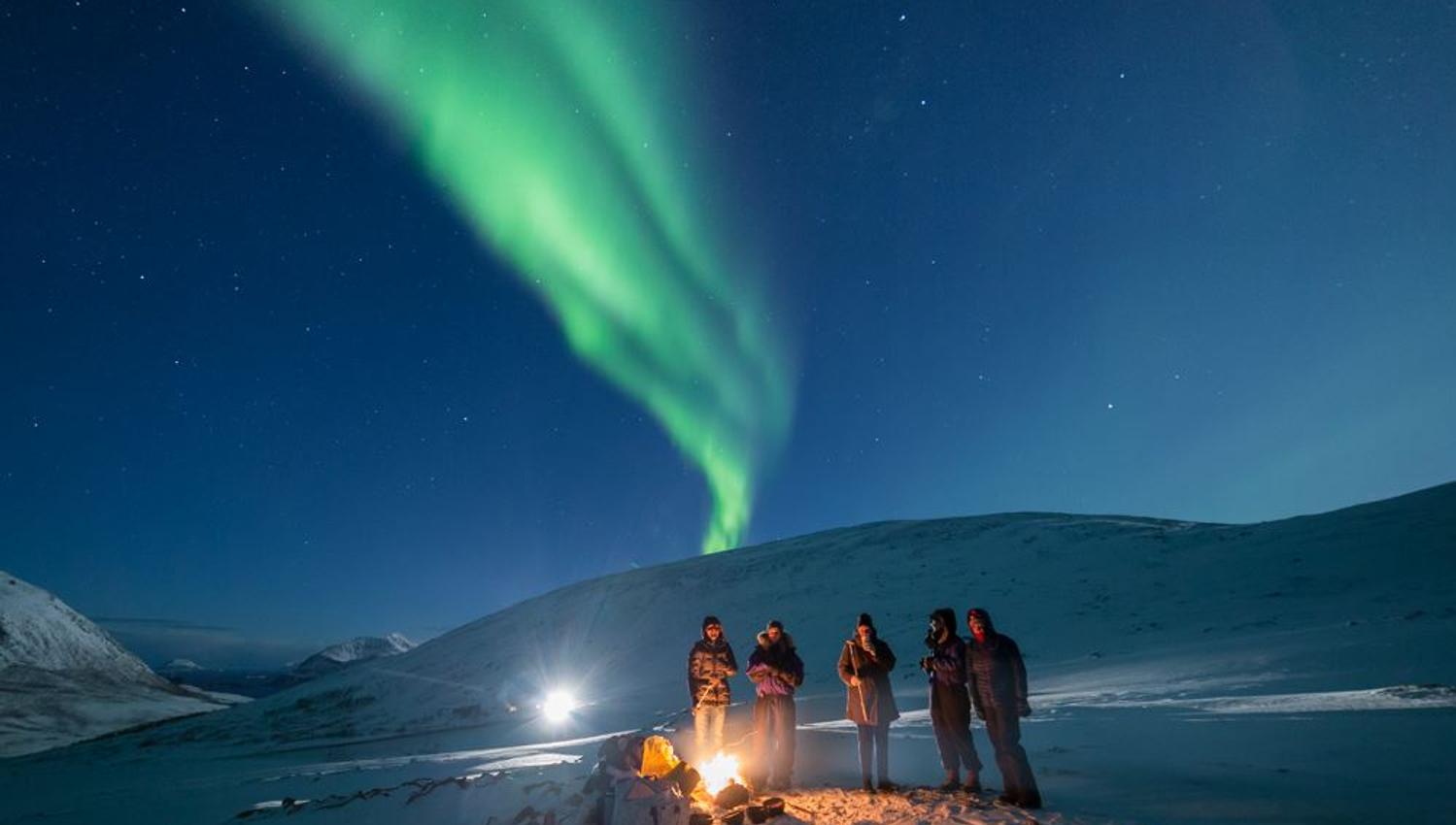 Guests surround the campfire while the Northern Lights fly by overhead