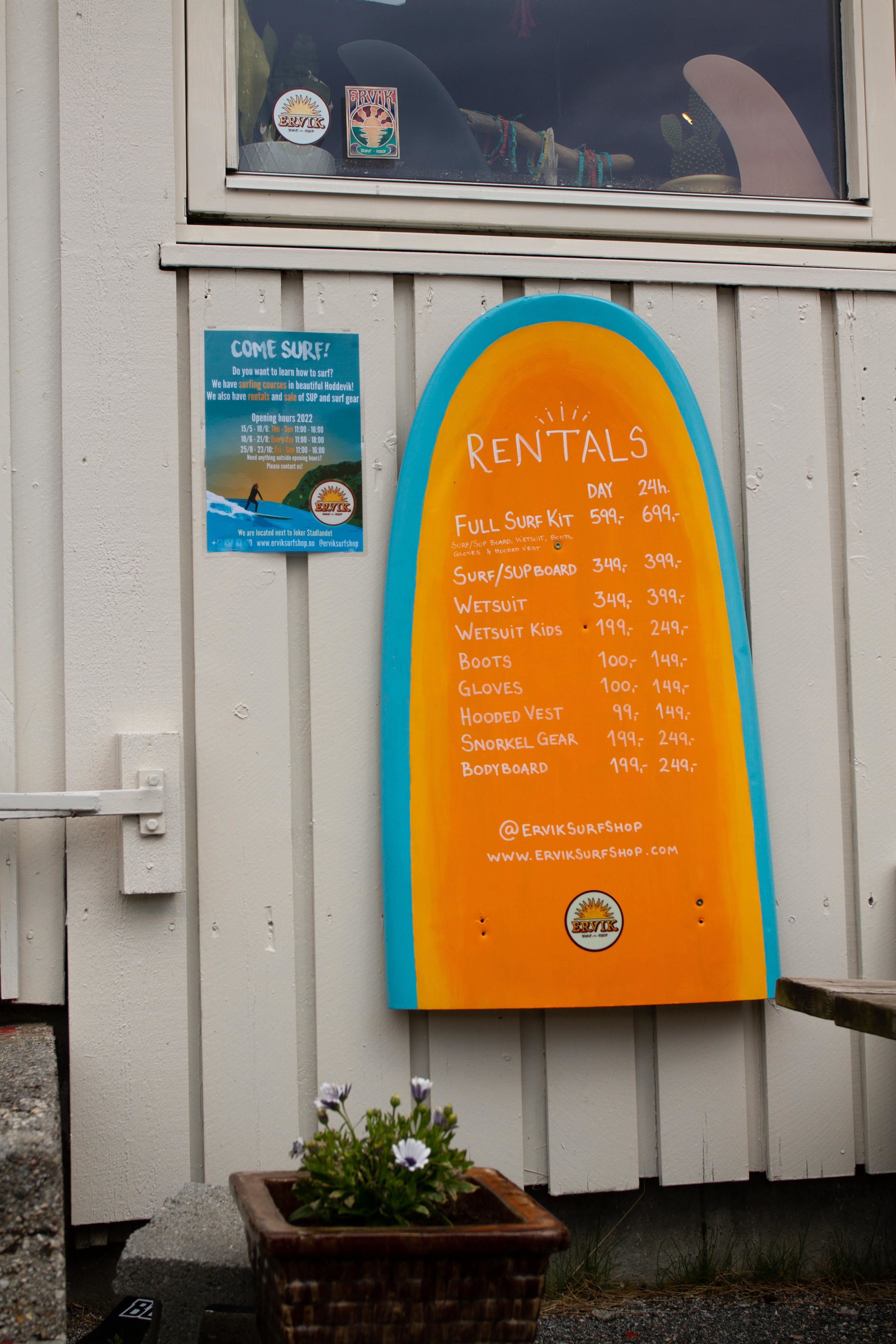 Eit oransje surfebrettforma skilt med utleigeprisar heng på ein lys vegg ved eit vindauge og ein plakat. ved Ervik Surfshop.