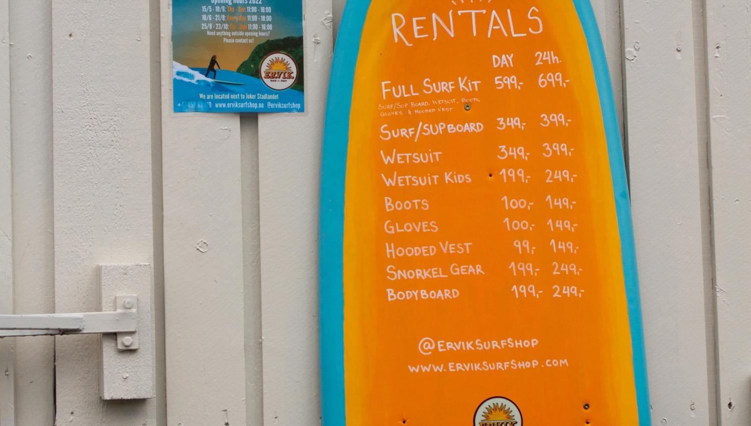 Eit oransje surfebrettforma skilt med utleigeprisar heng på ein lys vegg ved eit vindauge og ein plakat. ved Ervik Surfshop.