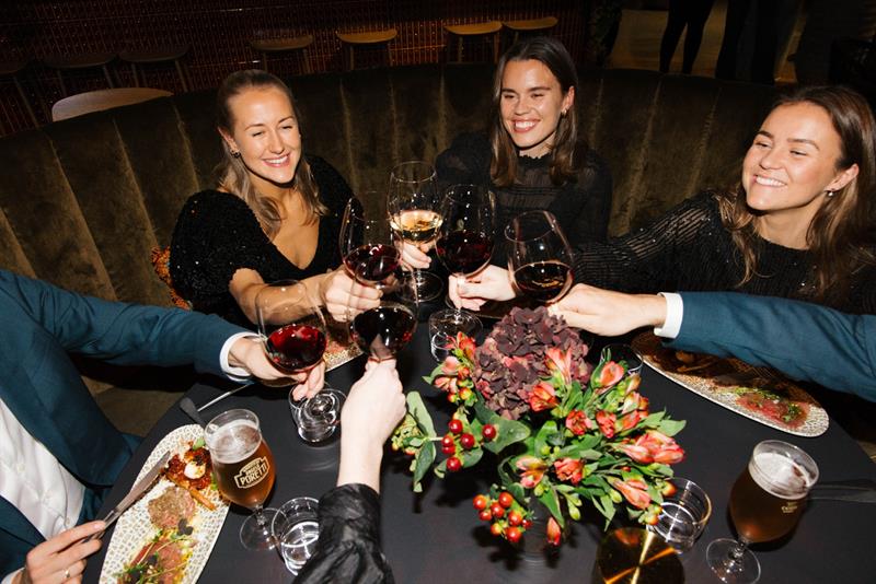 En gruppe mennesker skåler med vinglass over et restaurantbord, smilende og feststemte.