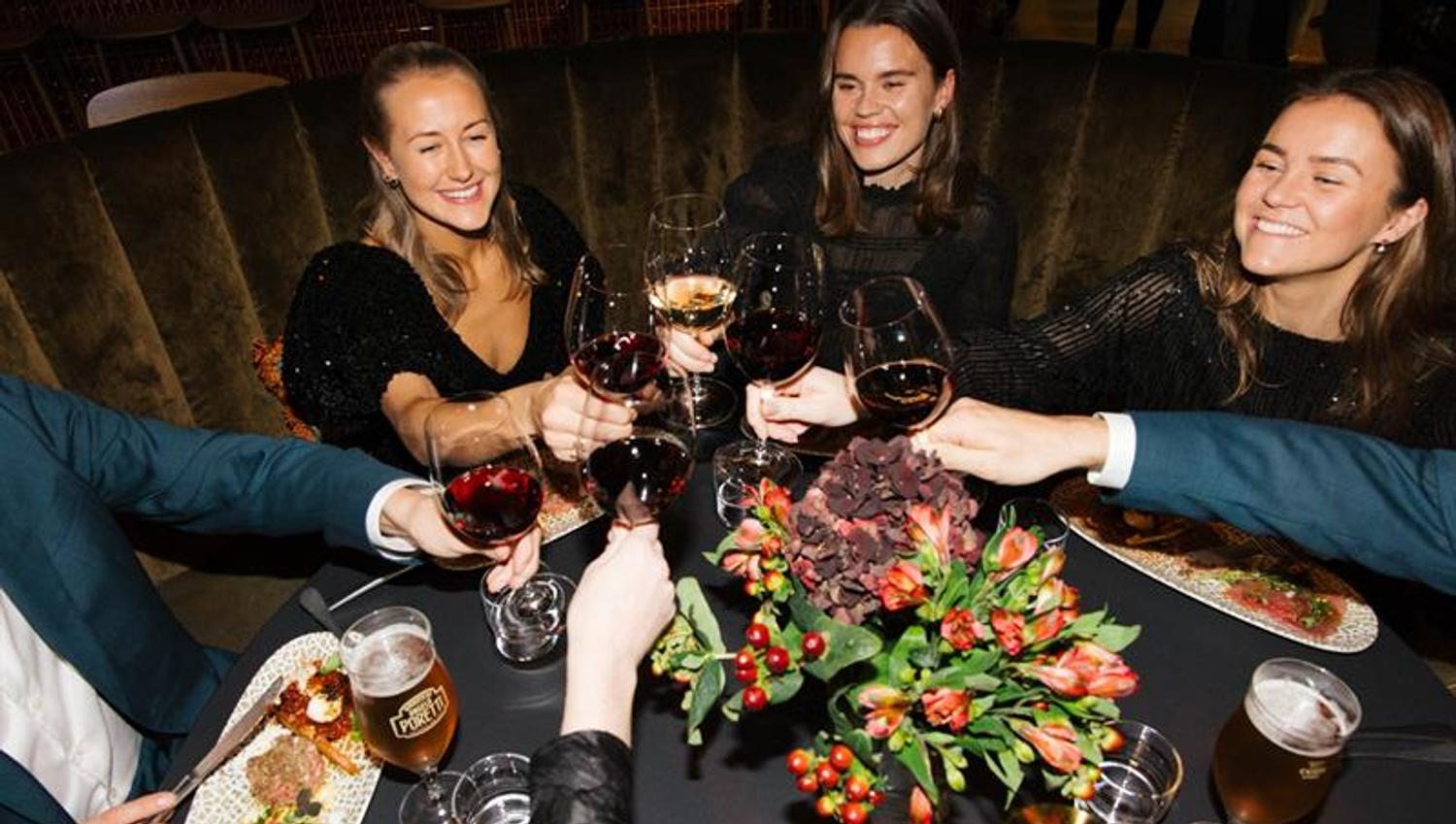En gruppe mennesker skåler med vinglass over et restaurantbord, smilende og feststemte.