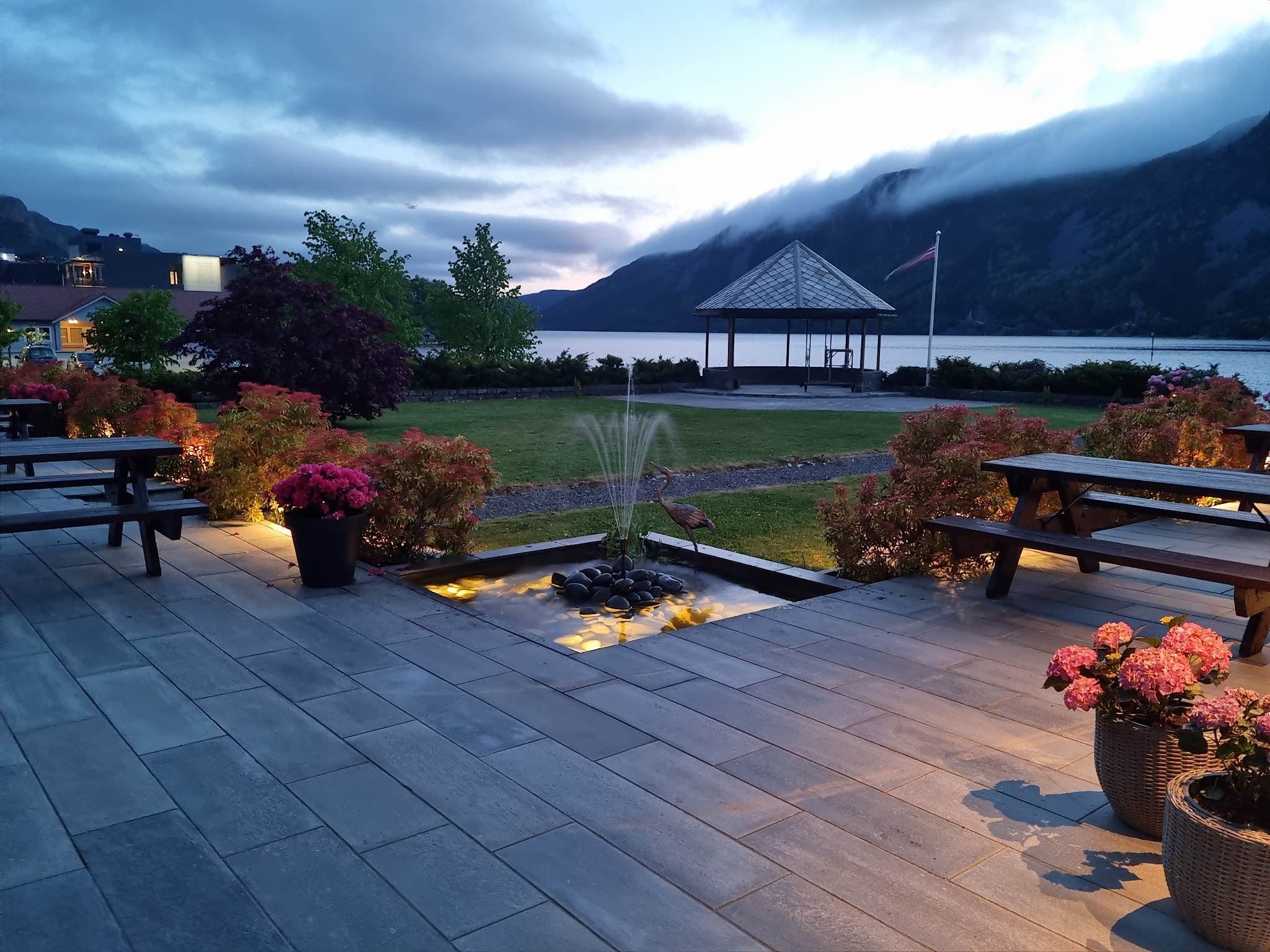 Uteplass med benkar, blomsterkrukker og liten fontene, med paviljong ved fjord og fjell i skumring..