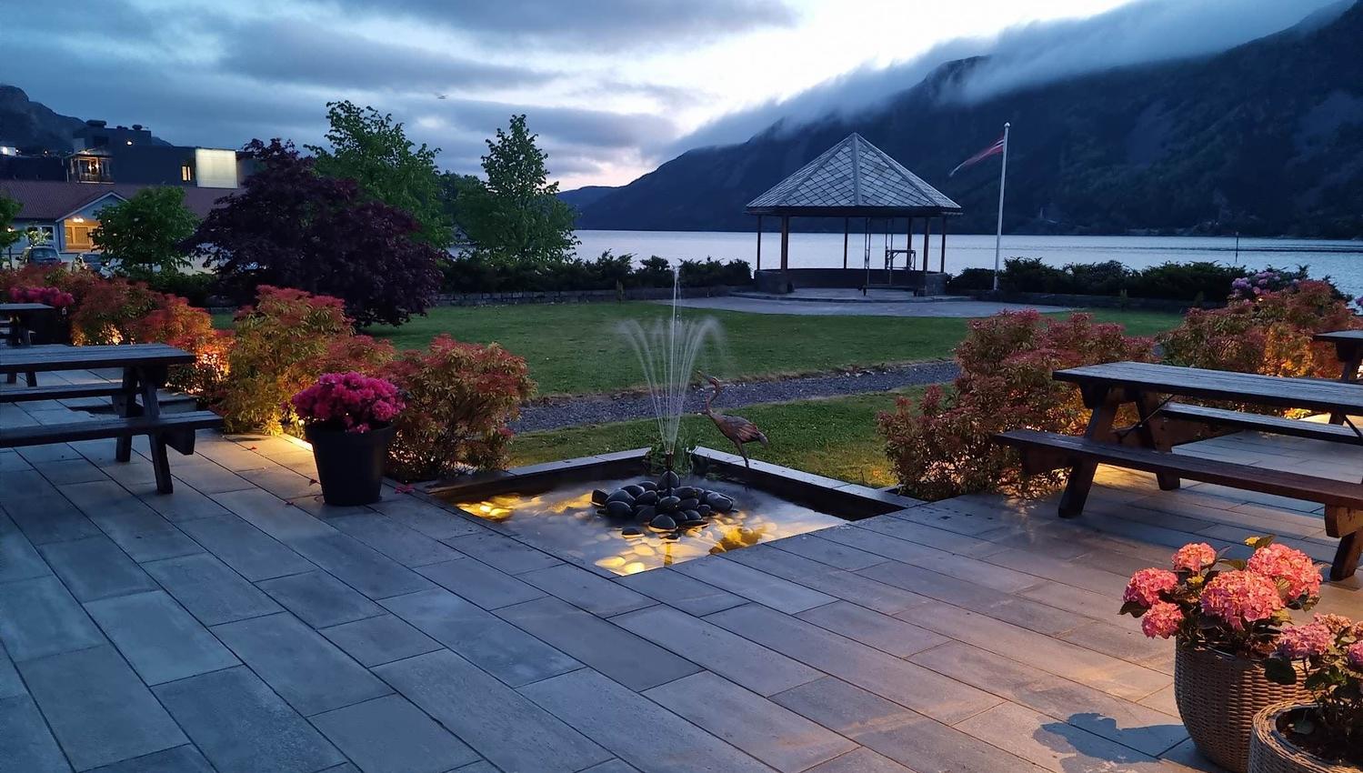 Uteplass med benkar, blomsterkrukker og liten fontene, med paviljong ved fjord og fjell i skumring..