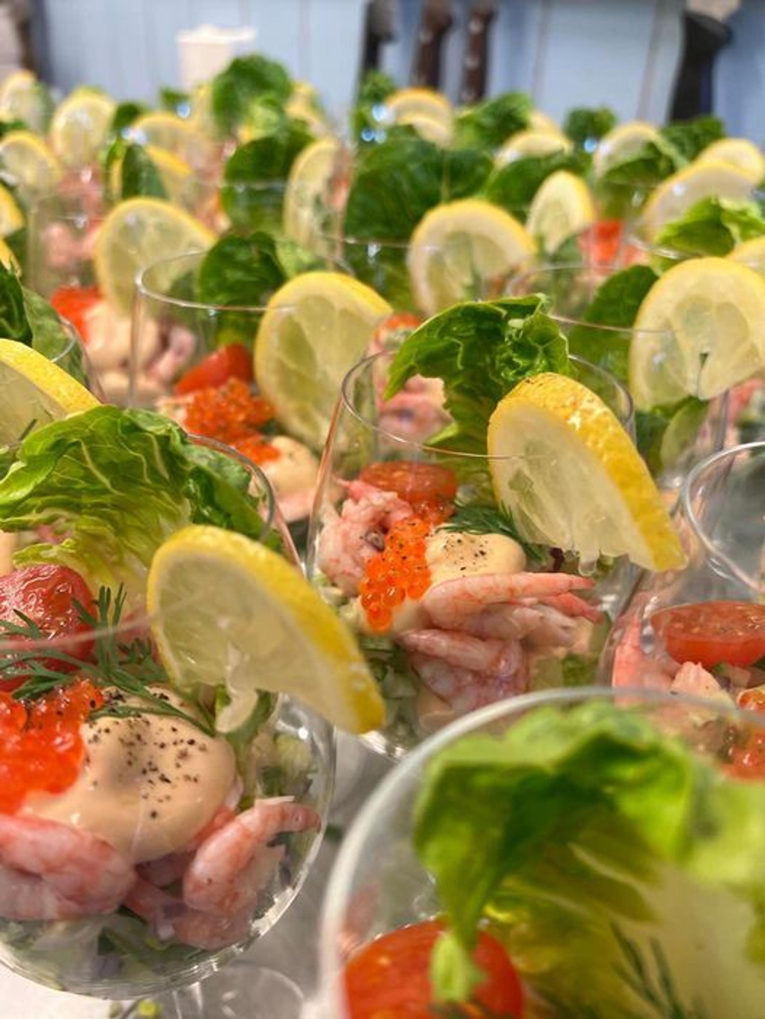Rows of clear appetizer cups filled with shrimp, sauce, roe, herbs, and lemon wedges with lettuce garnish.
