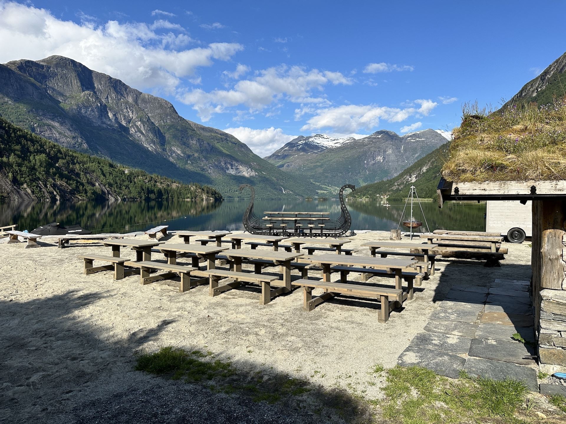 Wooden picnic tables on a sandy lakeshore with mountains and snowcapped peaks reflected in calm water under blue sky..