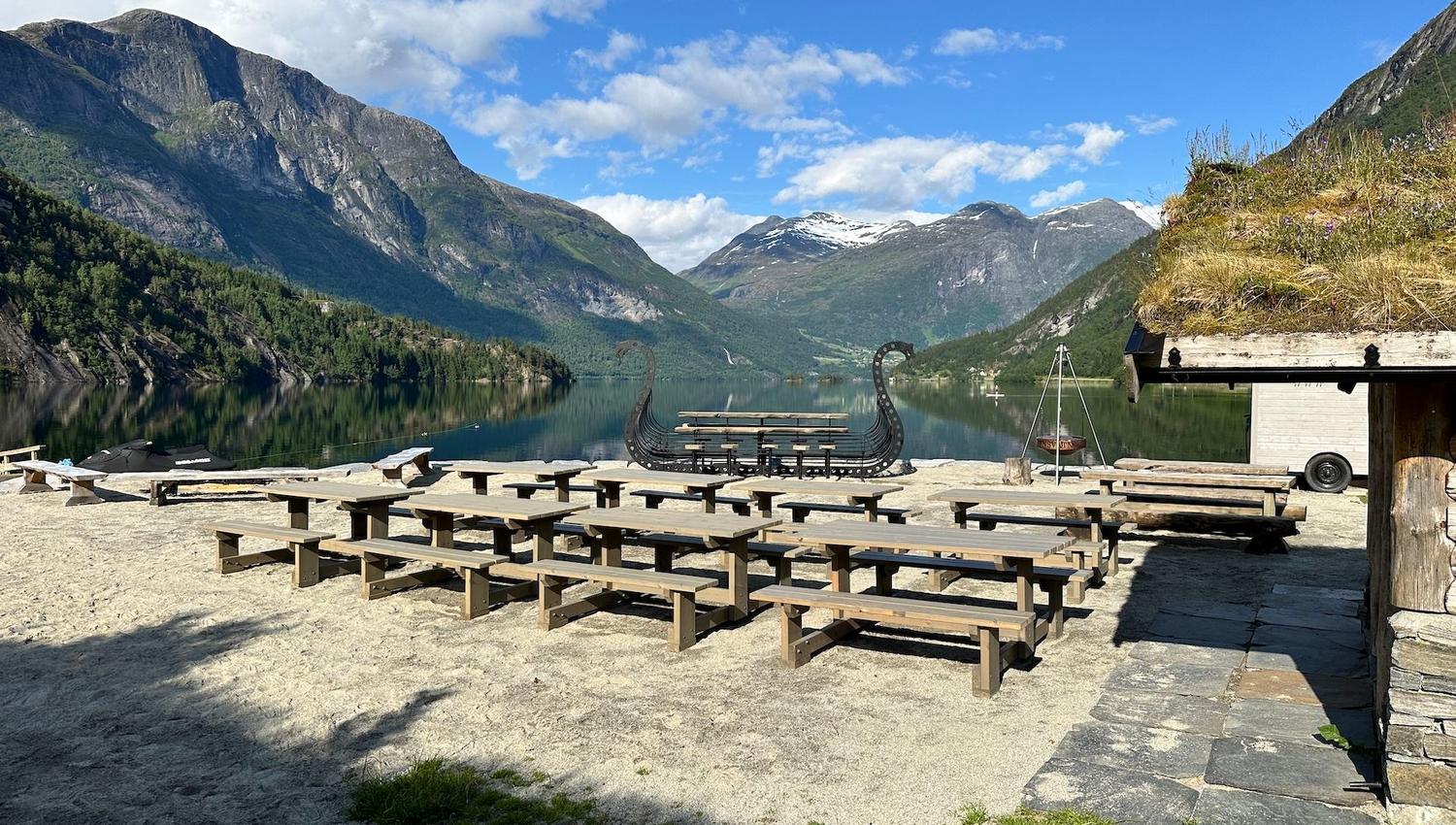 Wooden picnic tables on a sandy lakeshore with mountains and snowcapped peaks reflected in calm water under blue sky..