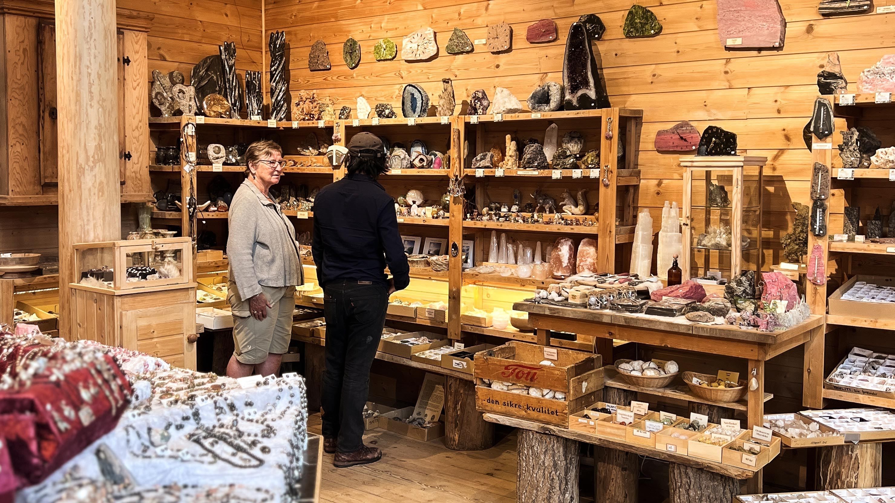 Two people browse wooden shelves filled with rocks, crystals, and gemstones in a warm, rustic shop..