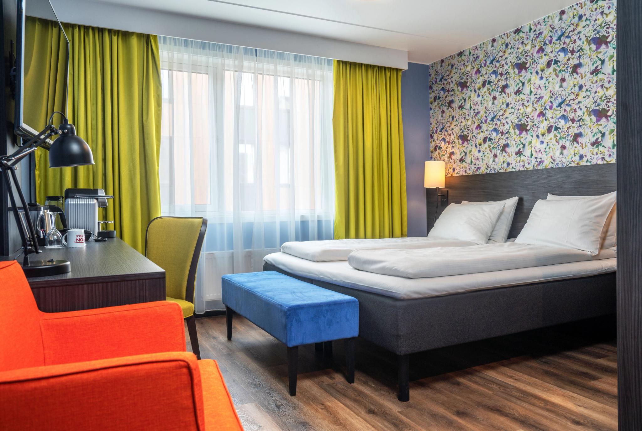Modern hotel room with a double bed, colourful decor, and a large window.